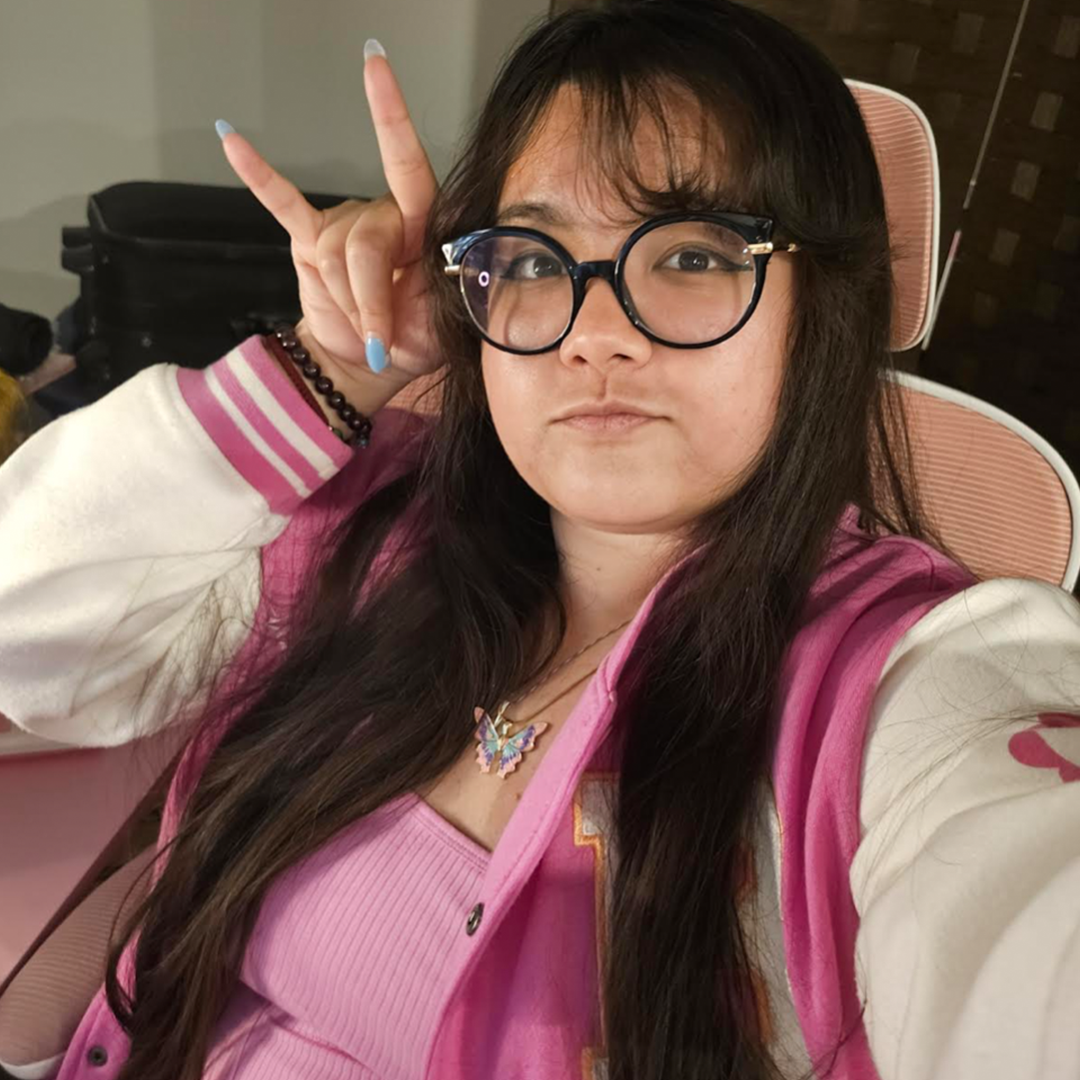 Close-up of Jessie Lo wearing a pink top with a white and pink jacket on top, a butterfly necklace, black glasses, and blue nailpolish. Jessie also has long dark brown hair.