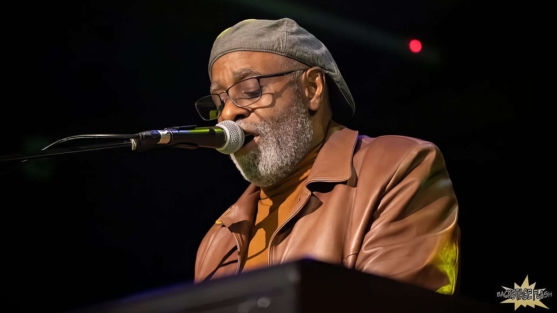 Phil Donaldson, wearing a cap and brown leather jacket, is singing or speaking into a microphone onstage at "The Last Waltz (Again)" in 2025.