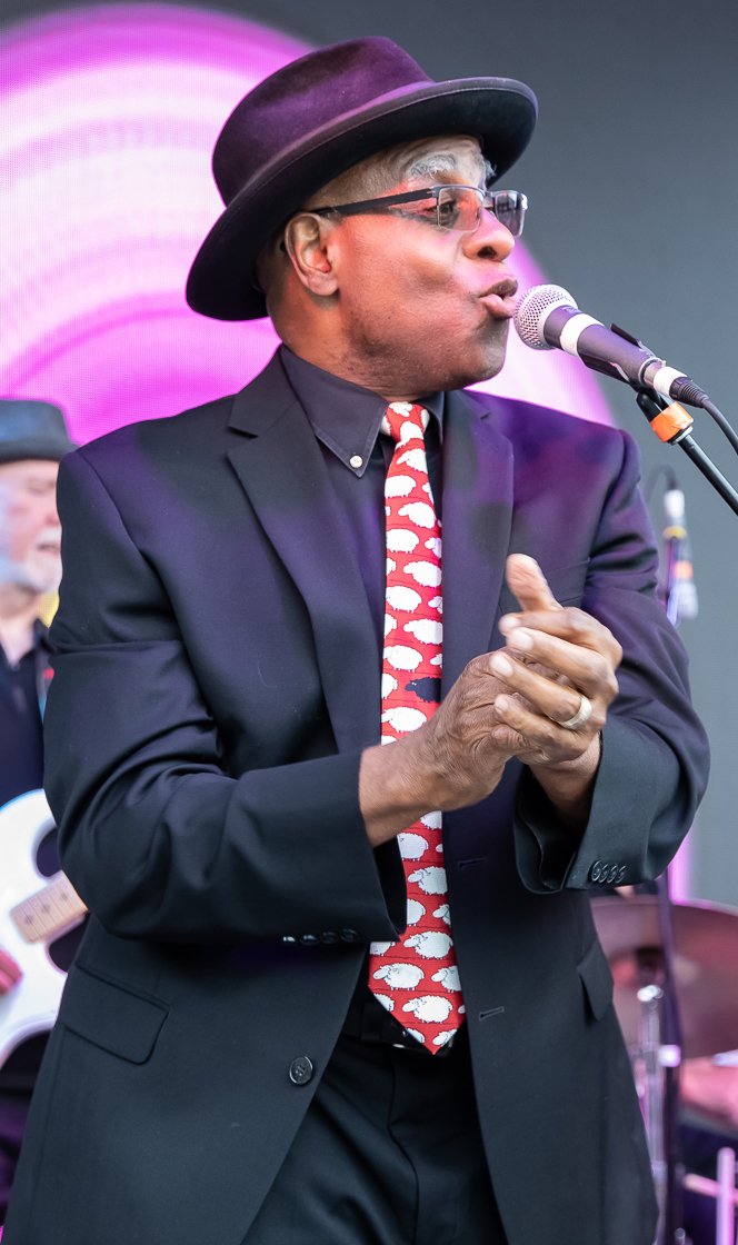 Phil Donaldson sings into a microphone onstage, wearing a black suit, wide-brimmed hat, sunglasses, and a red patterned tie.