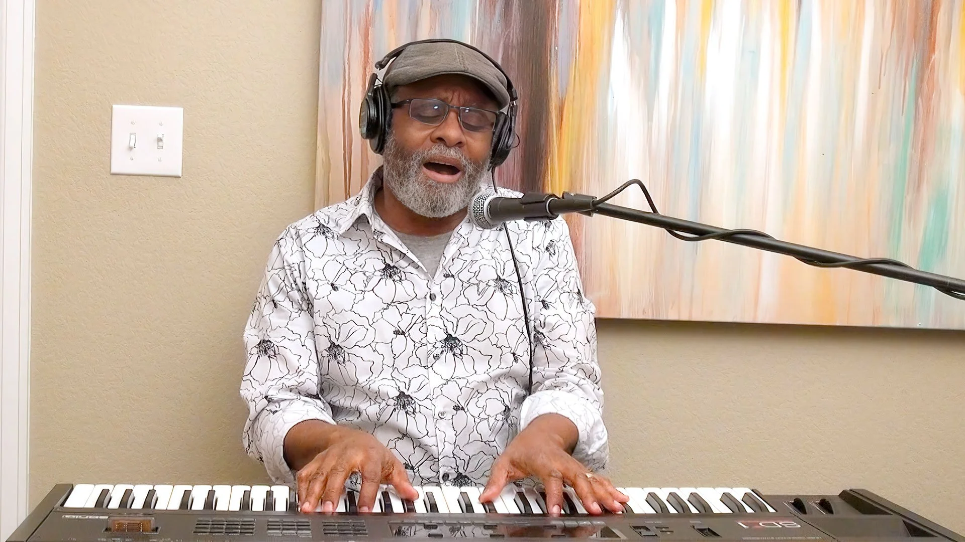 A bearded Phil Donaldson is singing into a microphone while playing a keyboard. He is wearing a gray cap and a white shirt with black floral patterns. Behind him is a colorful abstract painting on the wall.