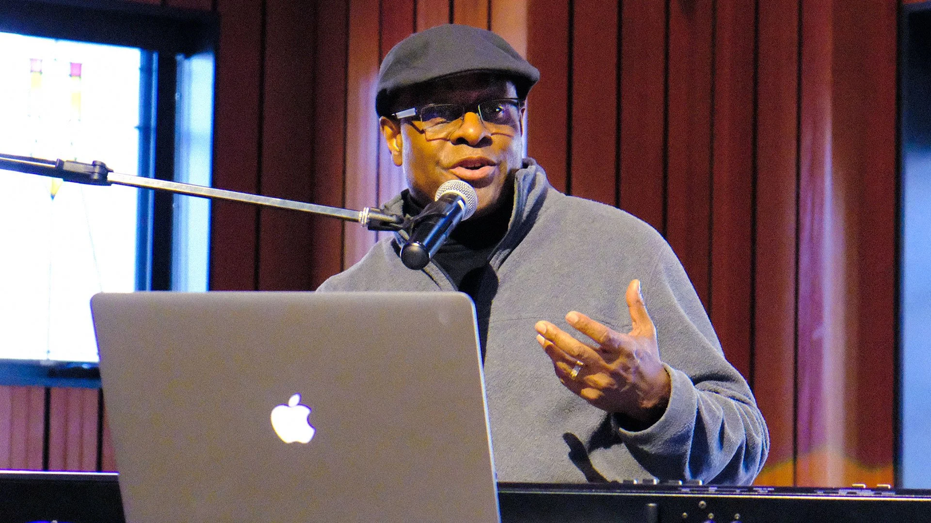 Phil Donaldson wearing a gray fleece, black cap, and glasses speaking into a microphone during a presentation, with a laptop and musical keyboard in front of him and wooden paneling in the background.