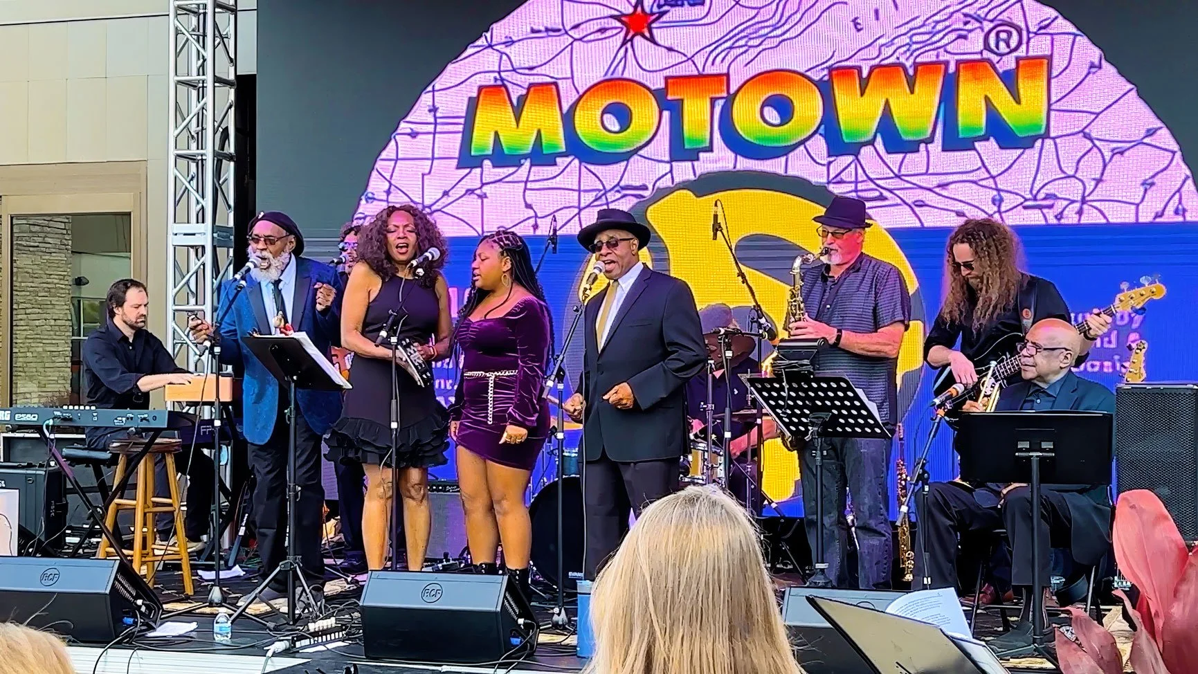 Phil Donaldson sings lead with FoCo Motown Revue on an outdoor stage, backed by vocalists, horns, keyboards, bass, and drums beneath a colorful Motown backdrop.