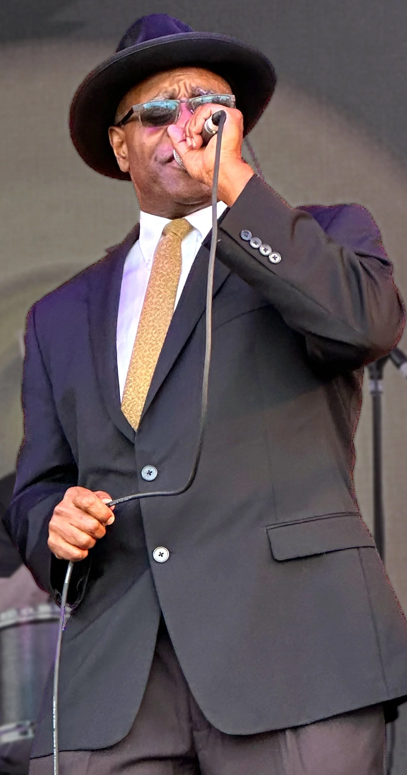 Close-up of Phil Donaldson singing powerfully into a handheld microphone, wearing a black suit, gold tie, wide-brimmed hat, and sunglasses.