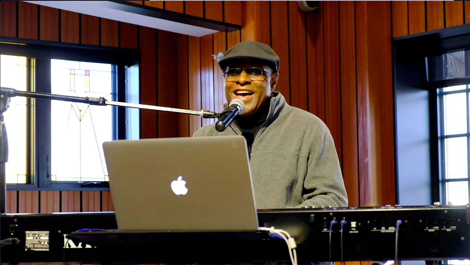 Phil Donaldson smiles while singing into a microphone at a keyboard, with a laptop open in a wood-paneled performance space.