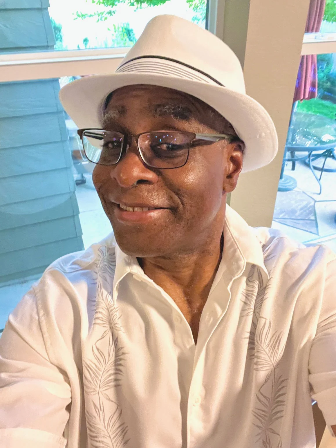 Phil Donaldson smiles for a casual indoor portrait, wearing glasses, a white fedora, and a white embroidered shirt.