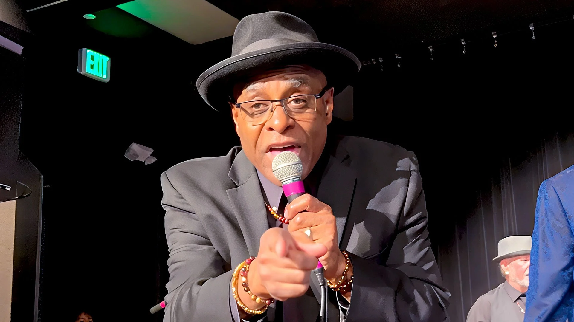 Closeup of Phil Donaldson singing and pointing to the camera. He is wearing a black suit and hat while holding a microphone. 