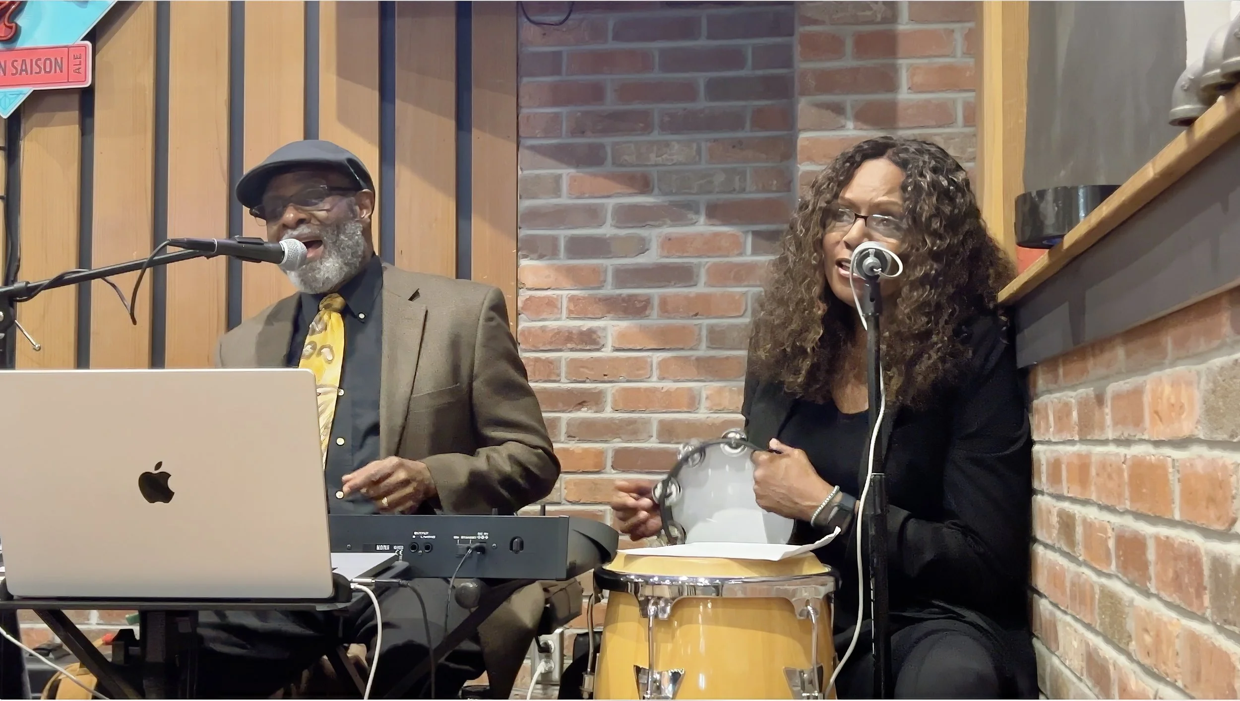 Two musicians performing indoors against a brick wall. The man on the left is singing into a microphone, wearing glasses, a hat, a suit, and a yellow patterned tie. He has a gray beard and is playing a laptop and a keyboard. The woman on the right is