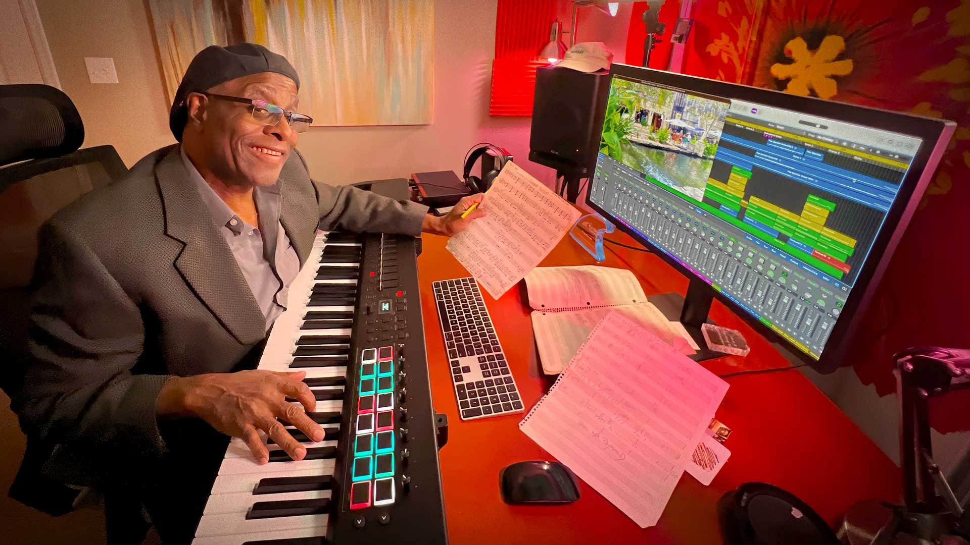 Phil Donaldson in his studio smiling at the keyboard while holding sheet music surrounded by recording gear and a digital workstation creating and arranging music with focus and joy.