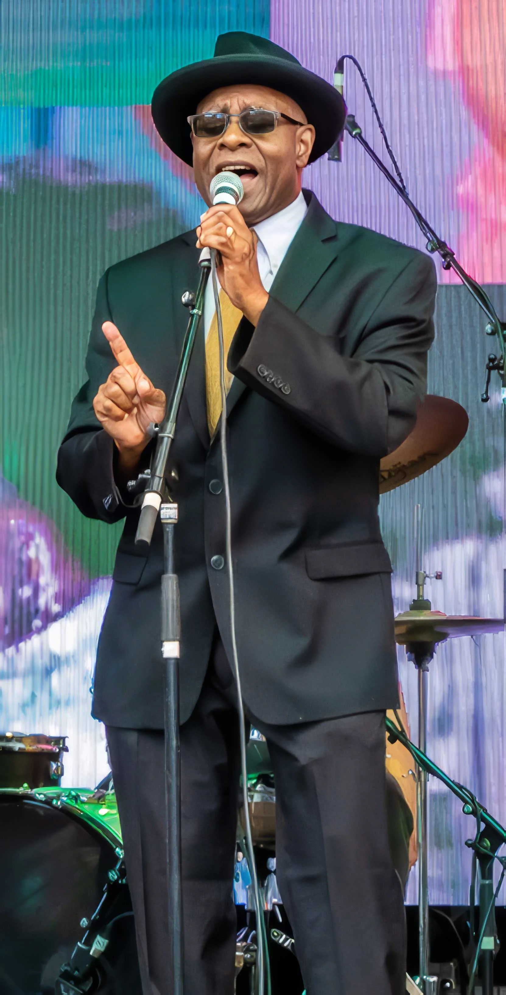 Phil Donaldson sings into a handheld microphone onstage, wearing a black suit, gold tie, wide-brimmed hat, and sunglasses.