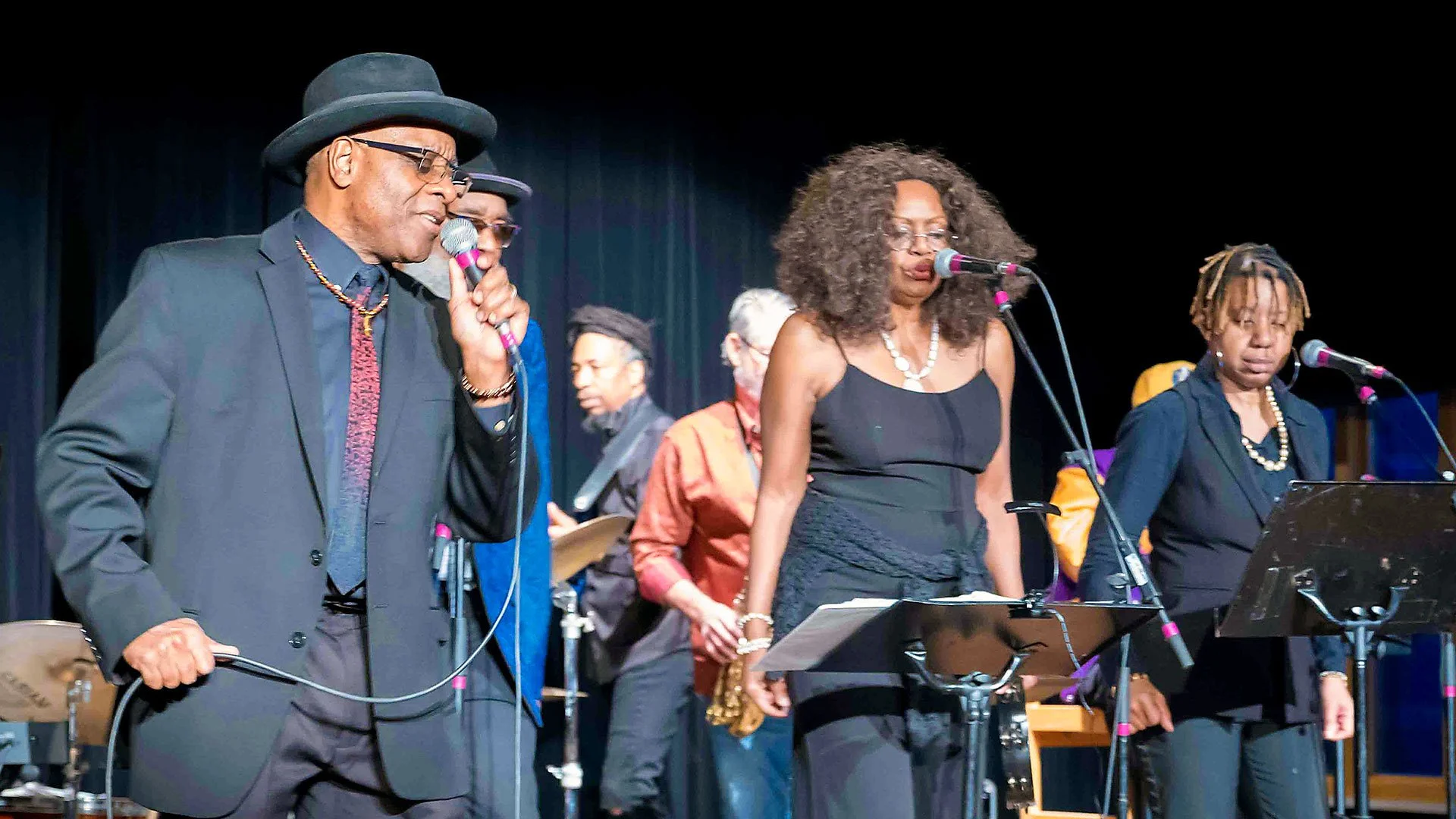Phil Donaldson singing with a group of six musicians performing on stage, three women and three men, with microphones, sheet music stands, and musical instruments in a concert setting.