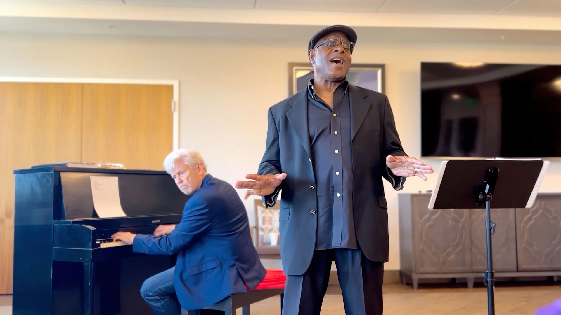 Phil Donaldson in a black blazer and newsboy cap singing with his hands gesturing, standing beside David Wohl, a man in a blue blazer playing a grand piano in a room with neutral-colored walls and a large television screen in the background.