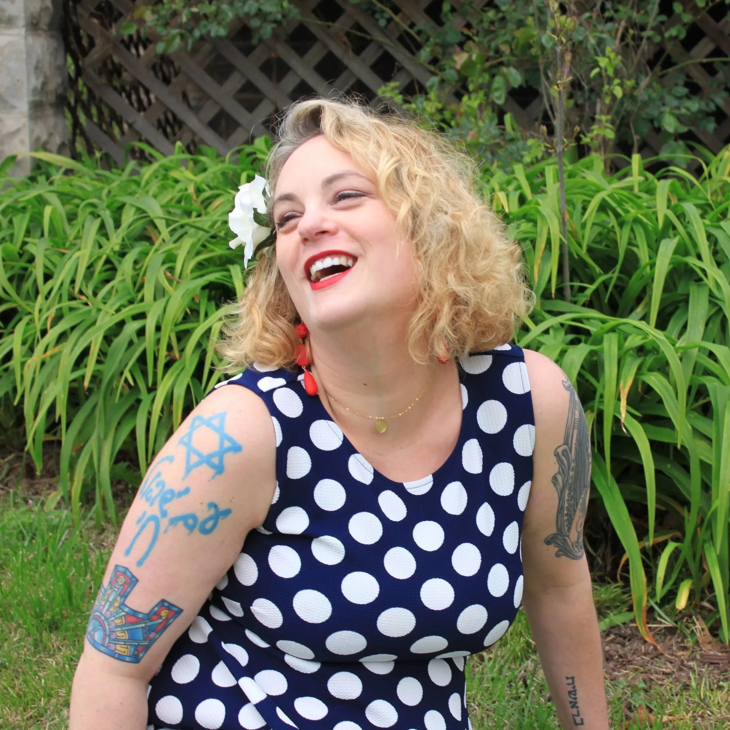 A woman with curly blonde hair, wearing a navy dress with white polka dots, and red lipstick, smiling in front of green plants and a wooden fence. She has a white flower in her hair and tattoos on her left arm.
