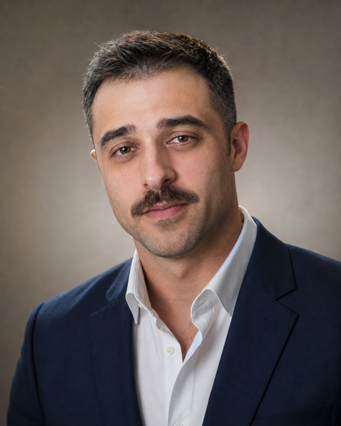 Headshot of a man with dark hair and a mustache, wearing a dark suit and white shirt, against a neutral background.