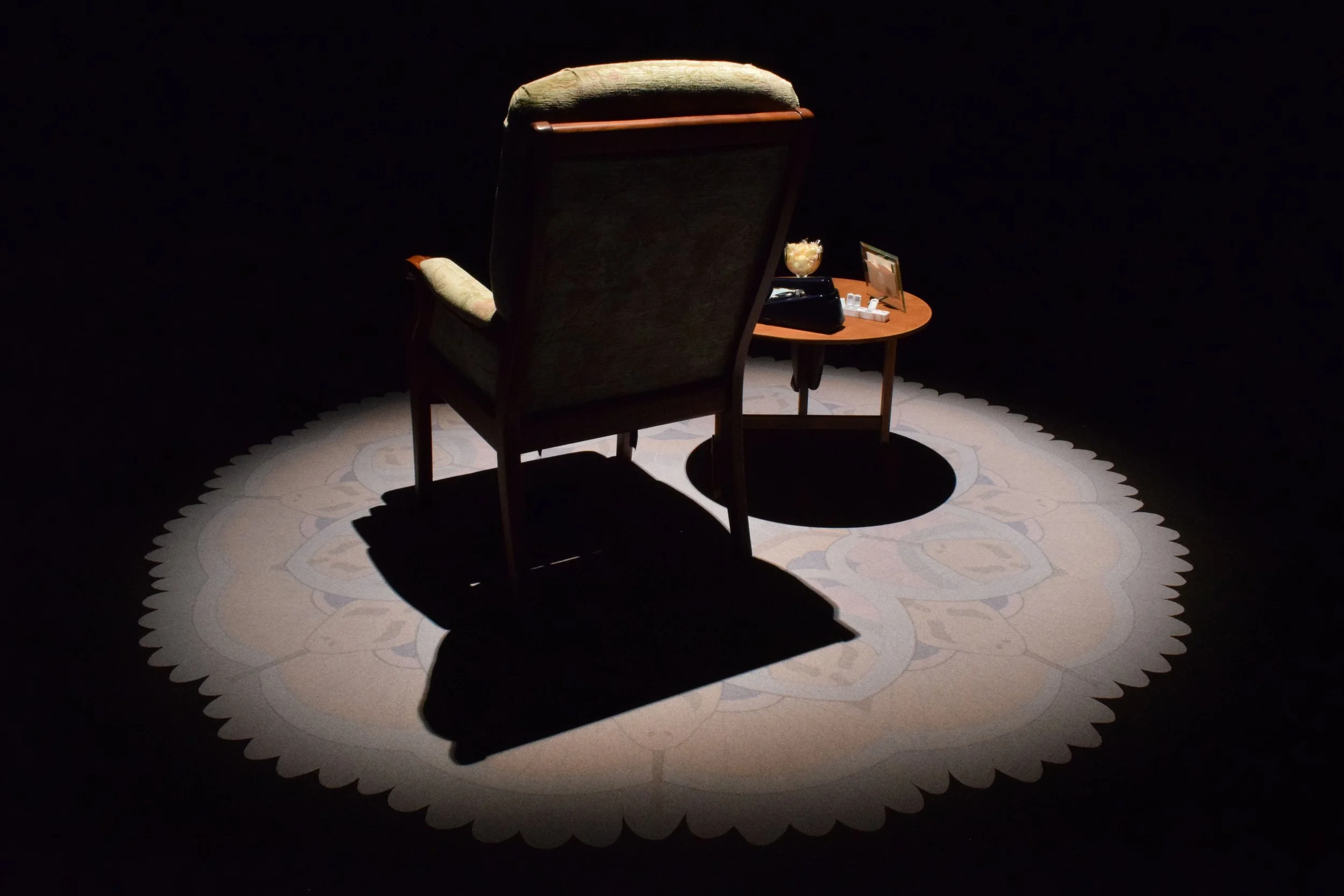A spotlight illuminating an armchair, a staged side table on top of a vinyl printed patterned rug forming part of an immersive installation.