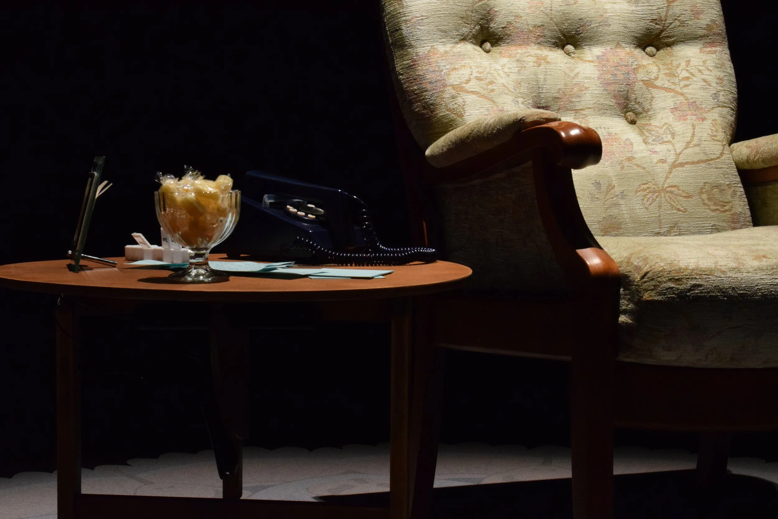 A wooden side table next to an arm chair with a glass bowl of boiled sweets, a vintage telephone, and a small framed picture as part of an immersive installation.
