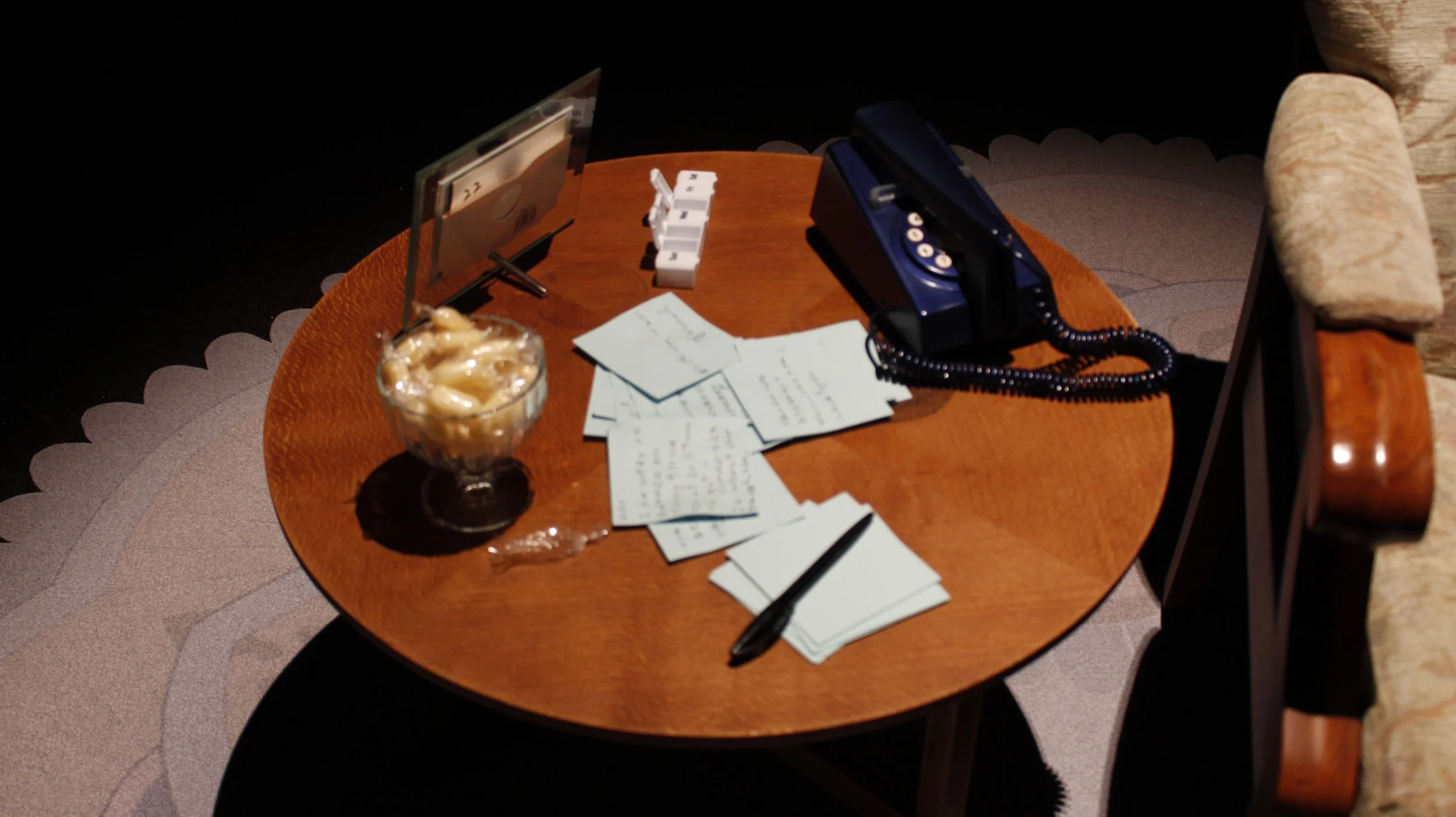 A wooden side table with a glass bowl of boiled sweets, scattered handwritten notes, a blue trim telephone, and a pill box as part of an immersive installtion.
