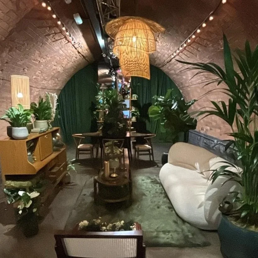 Immersive living-room setting filled with plants, furniture and warm lighting beneath brick arches.