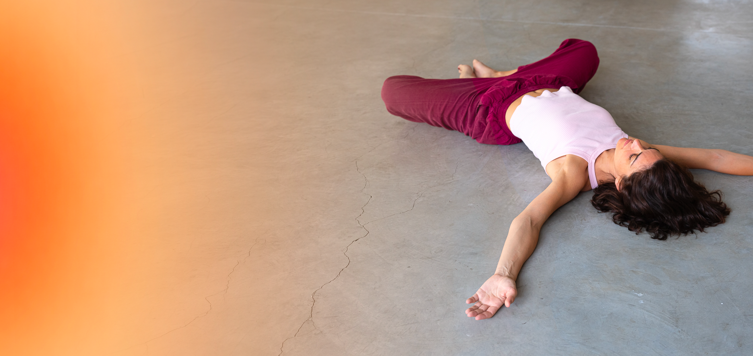 Une femme allongée sur un sol en béton, portant un t-shirt blanc et un pantalon marron, en position de relaxation, pratique le MUNZ FLOOR, yeux fermés.