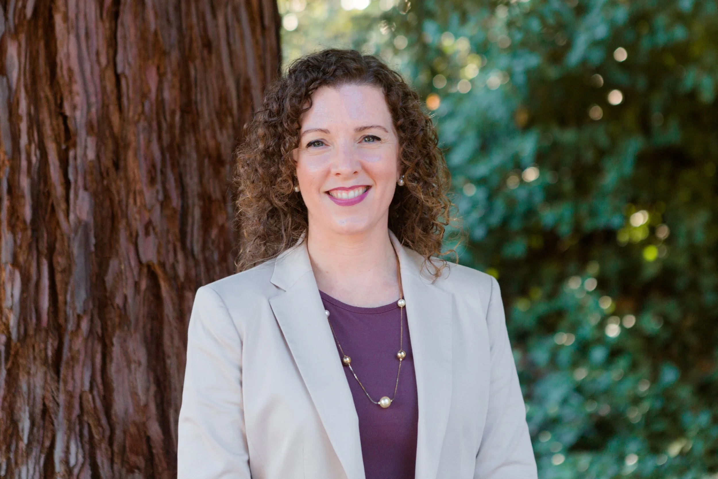 A woman with curly brown hair smiling outdoors next to a large tree with green foliage in the background.