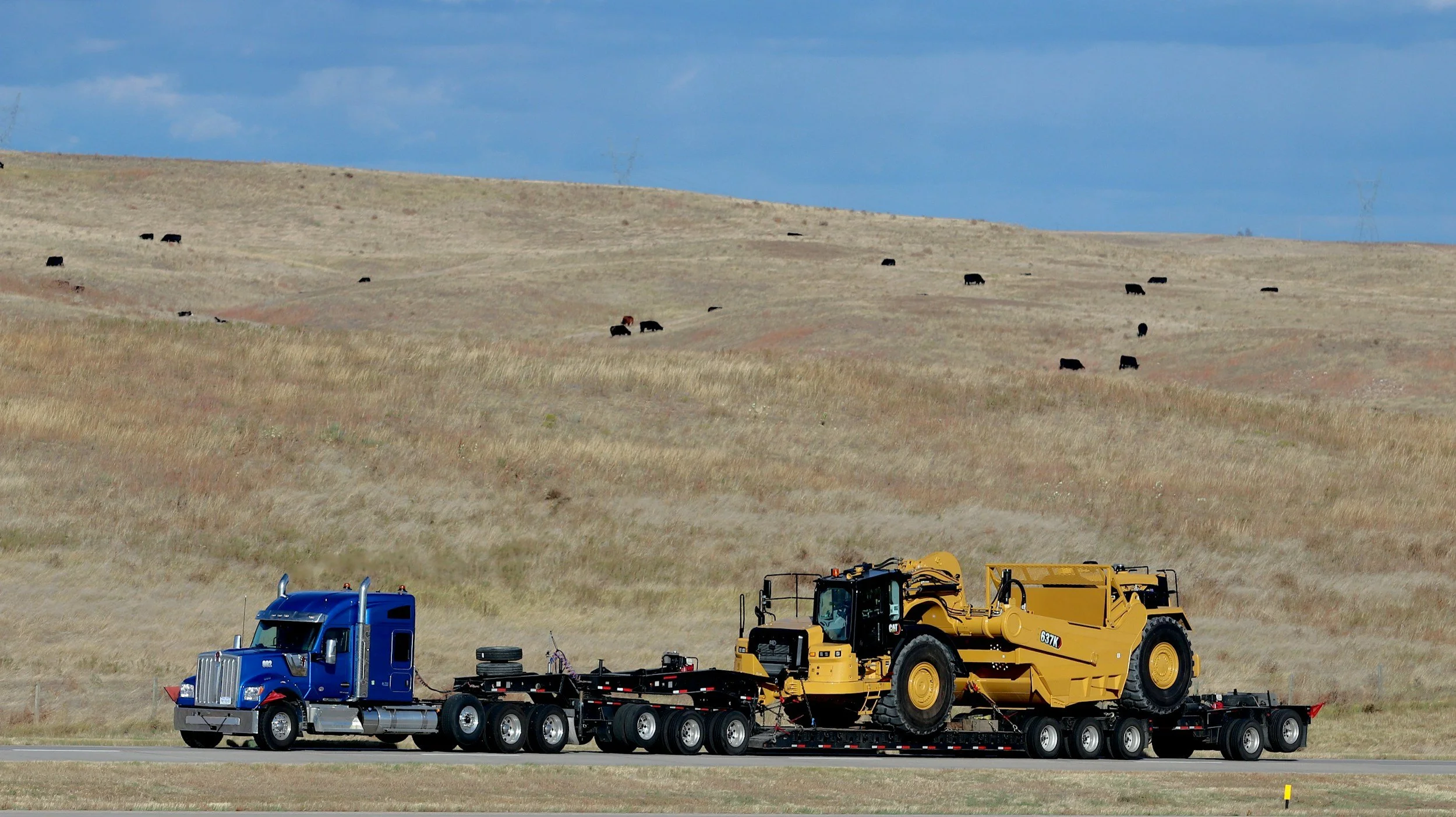 A large blue semi-truck transporting a yellow construction vehicle on a flatbed trailer through a rural landscape with rolling hills and sparse grass, under a partly cloudy sky.