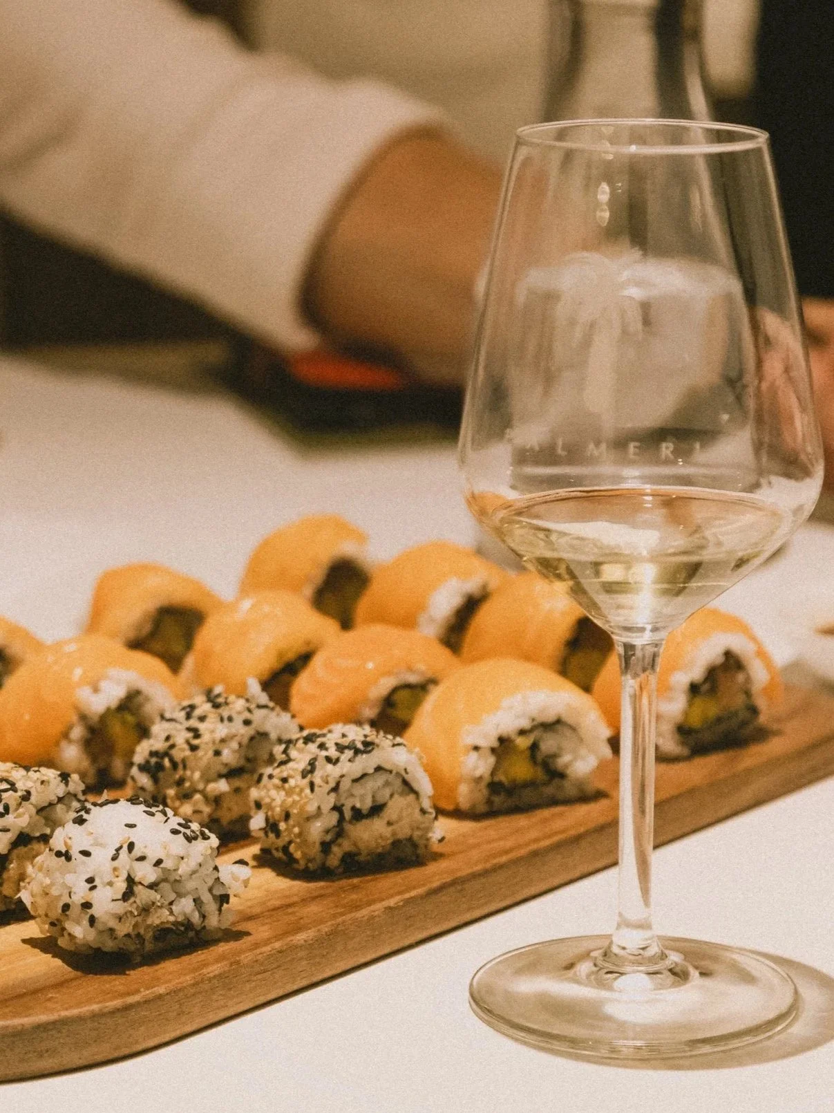 Verre de vin blanc devant un plateau de sushi, avec des rouleaux de sushi en arrière-plan.
