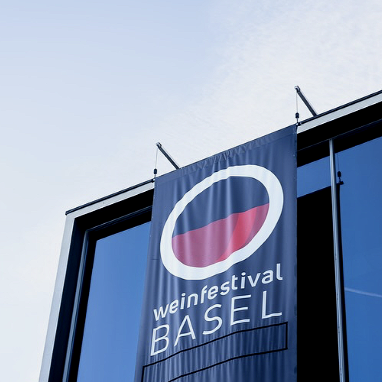Une grande bannière bleue suspendue à un bâtiment moderne en verre, proclamant le 'Weinfestival Basel' avec un logo circulaire coloré en rouge, blanc et bleu.
