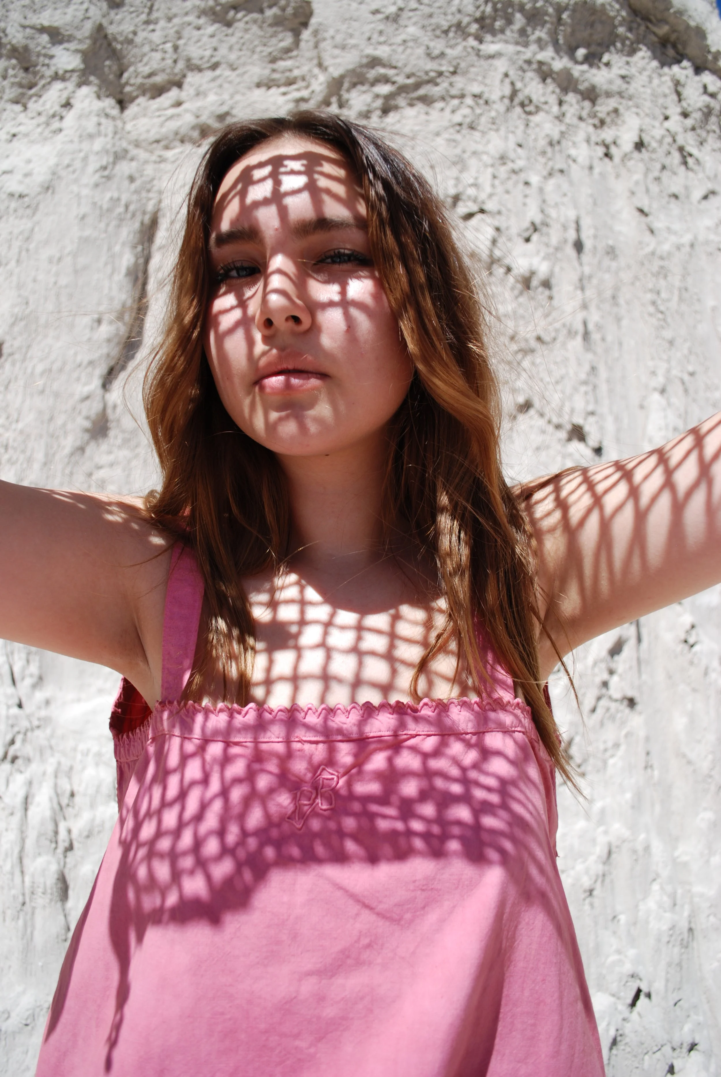 A young woman with wavy brown hair standing in front of a white textured wall, casting a shadow of a decorative metal lattice pattern onto her face and pink dress during sunlight.