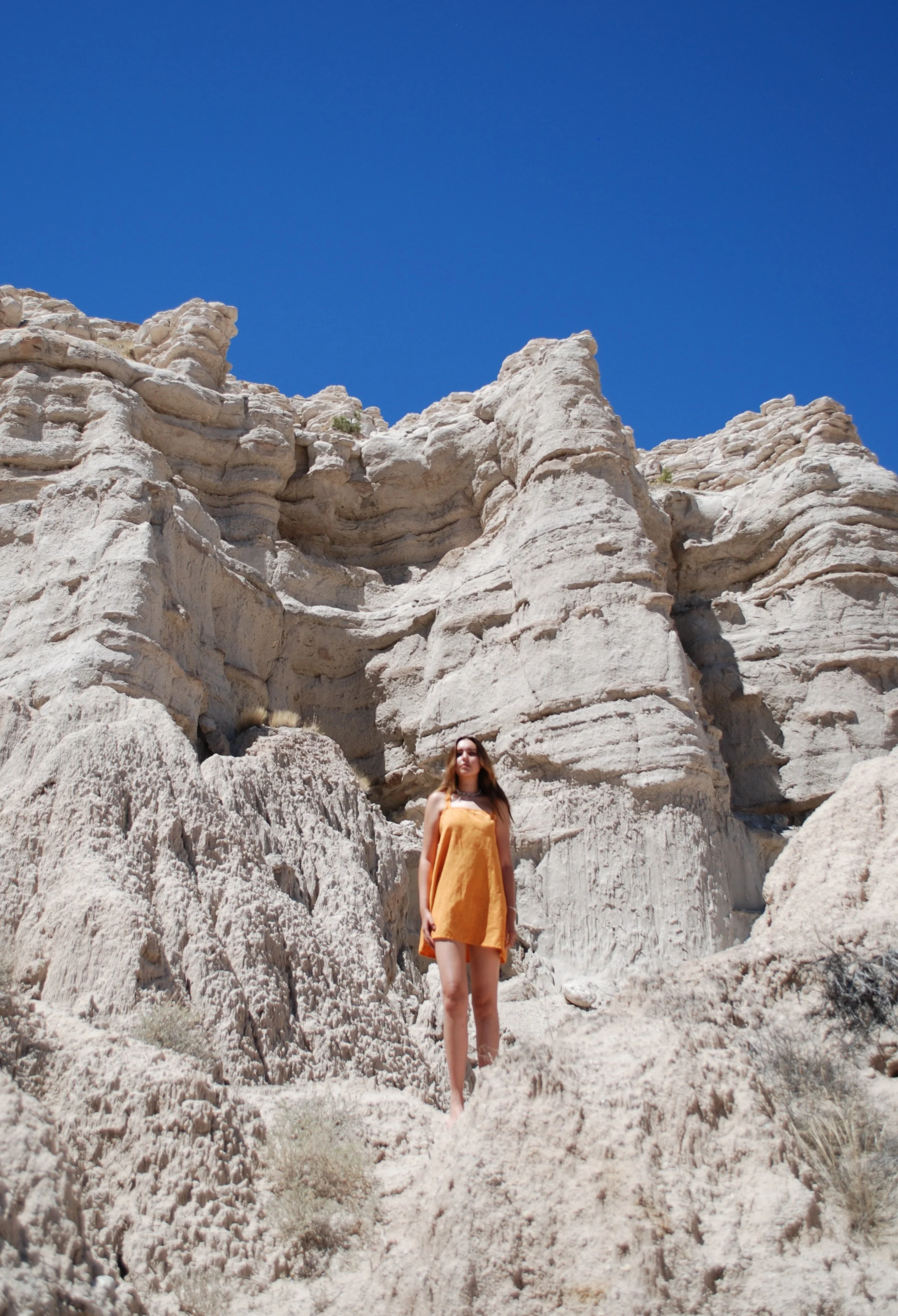 A woman in an orange dress walking on a rocky, white, desert landscape with large rock formations under a clear blue sky.