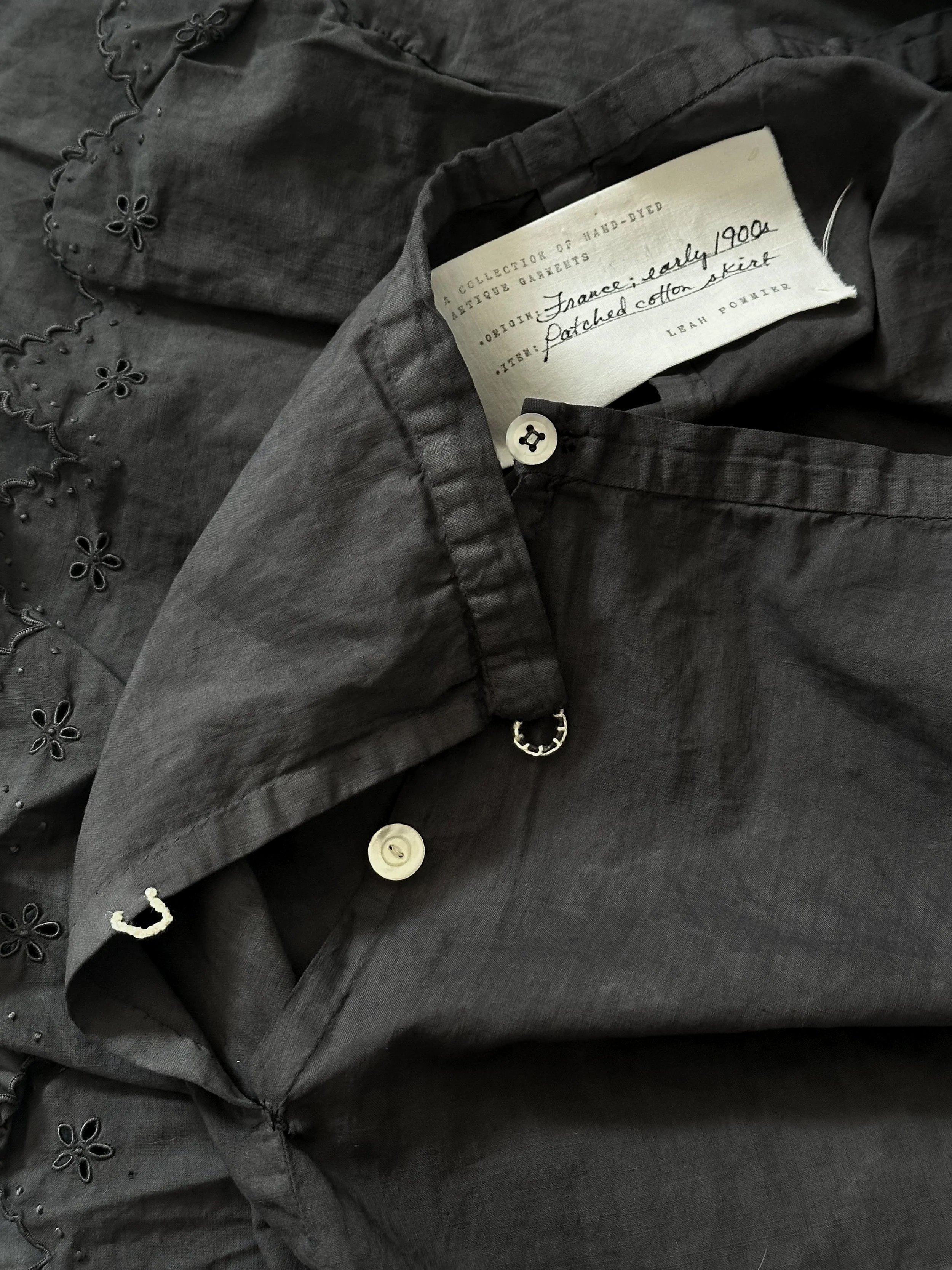 Close-up image of a dark gray vintage skirt with eyelet embroidery, showing buttons and a fabric label.
