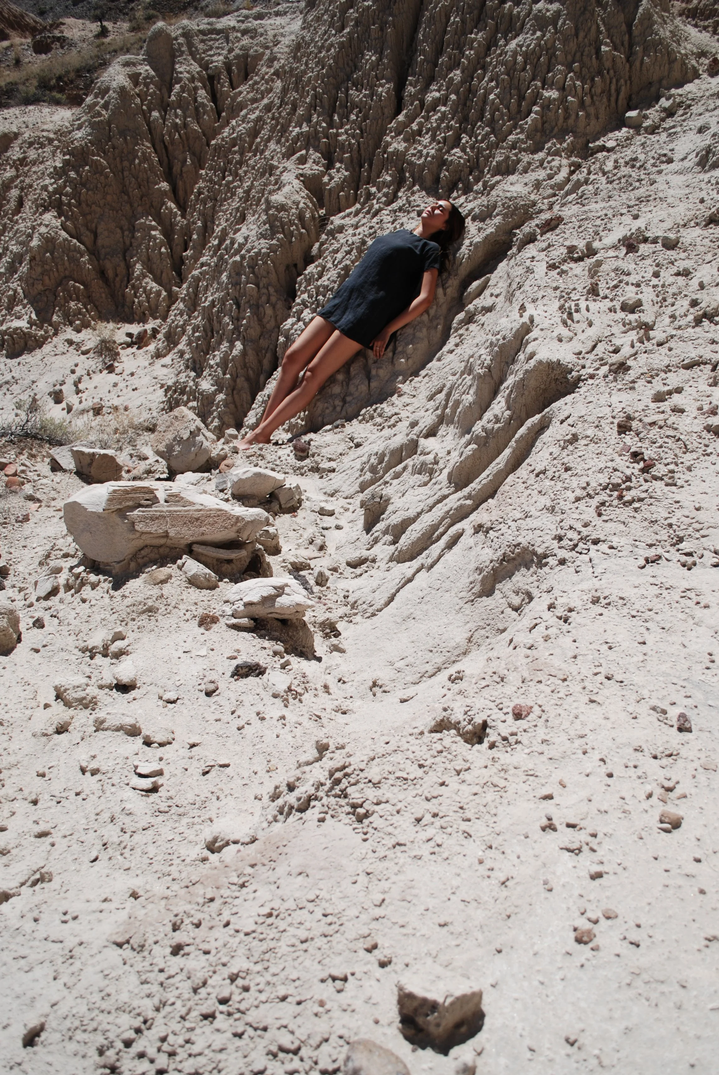 A woman lies on a rocky, desert landscape with eroded, beige-colored cliffs in the background, wearing a black dress with her eyes closed.
