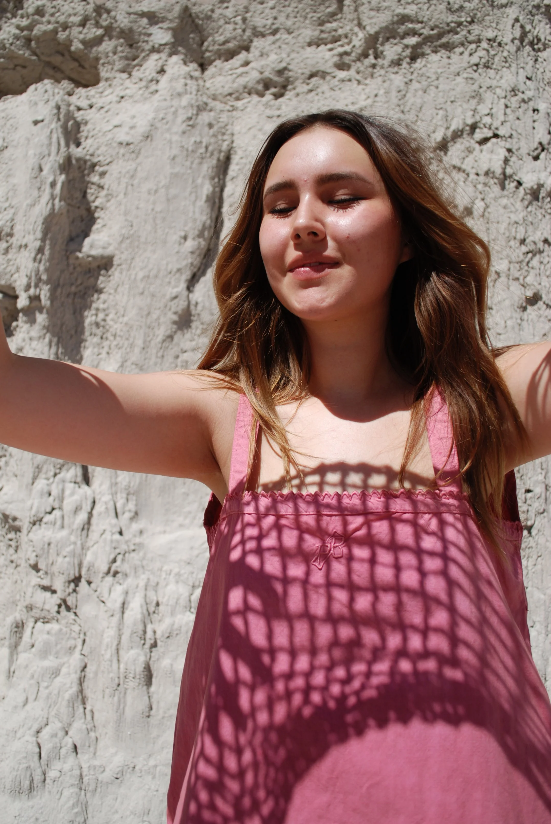 A young woman with brown hair and a pink sleeveless dress standing against a rocky, white cliff wall, with sunlight casting a shadow pattern on her dress.