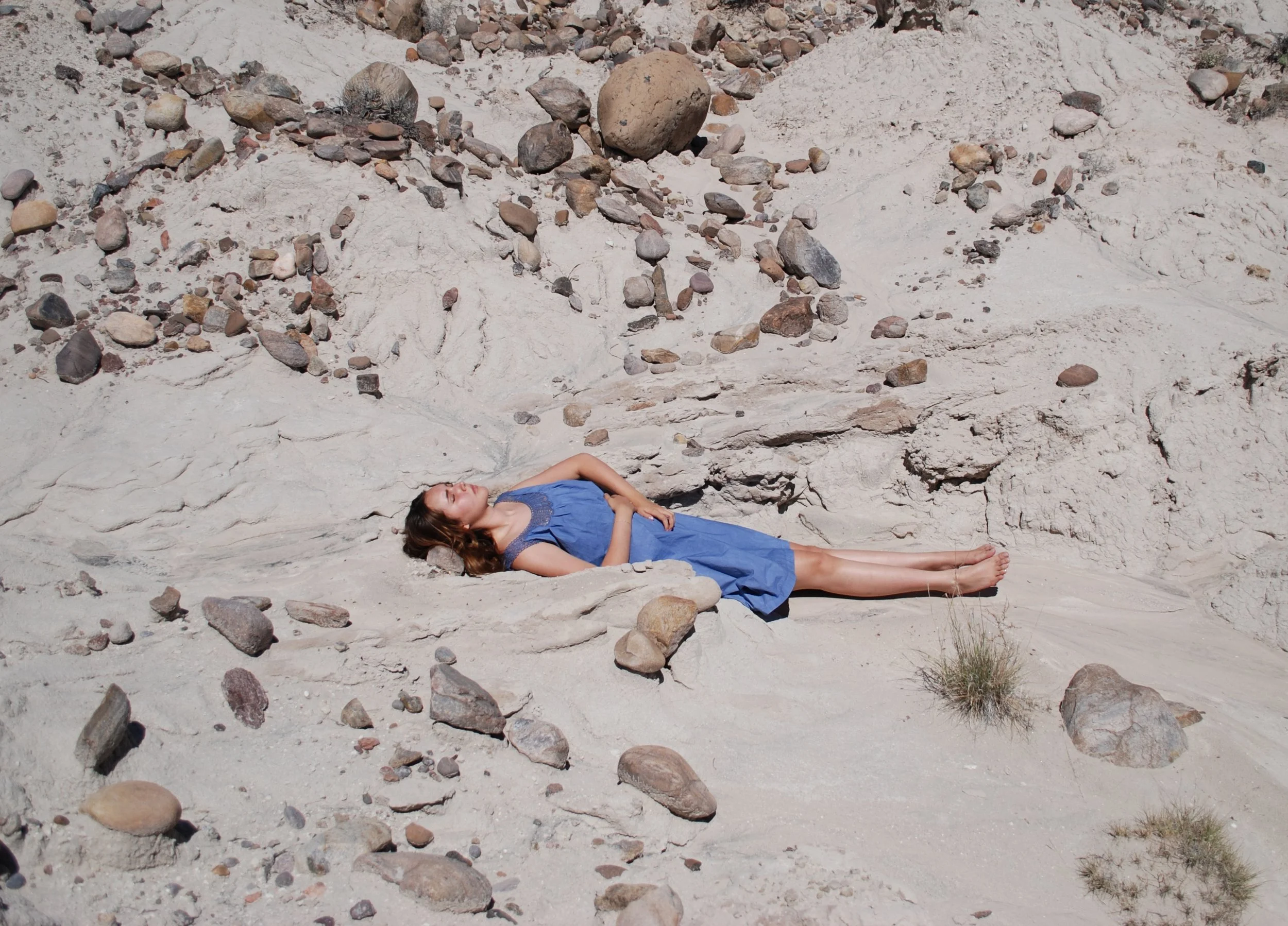 A woman in a blue dress is lying down on a sandy rocky landscape with her eyes closed, surrounded by various small and large rocks.