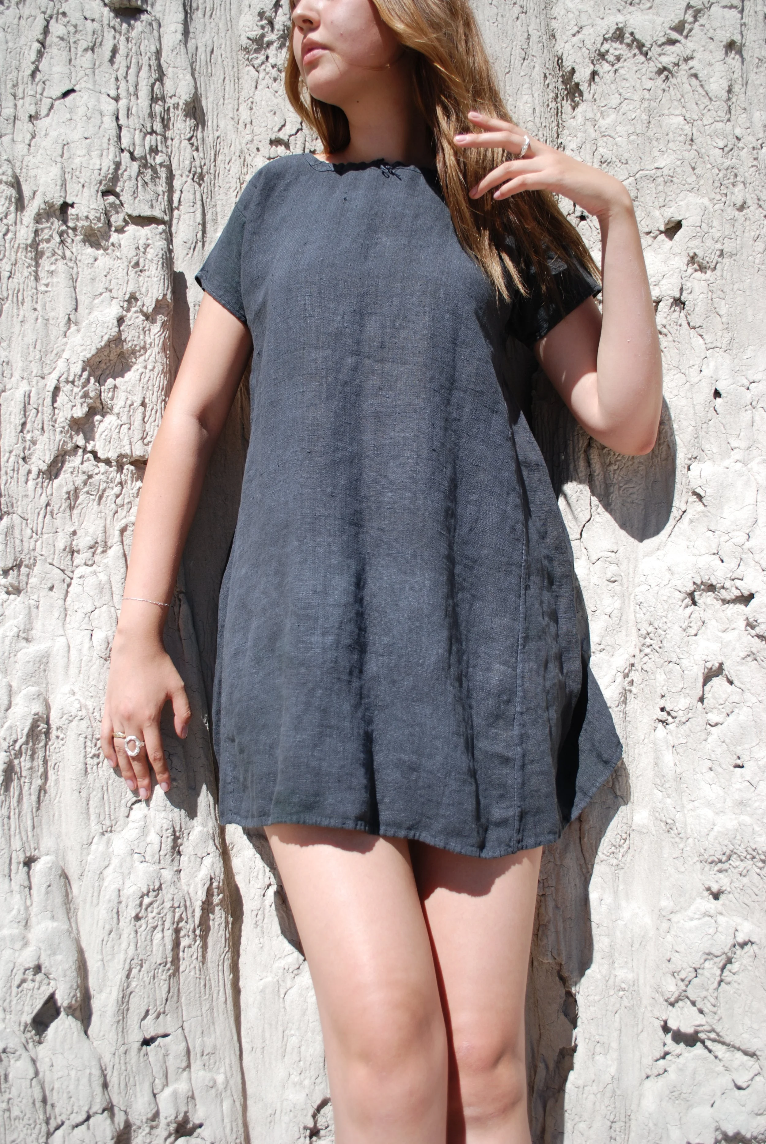 A woman with long light brown hair wearing a dark gray dress standing against a textured white stone wall, partially visible from her nose to her legs.