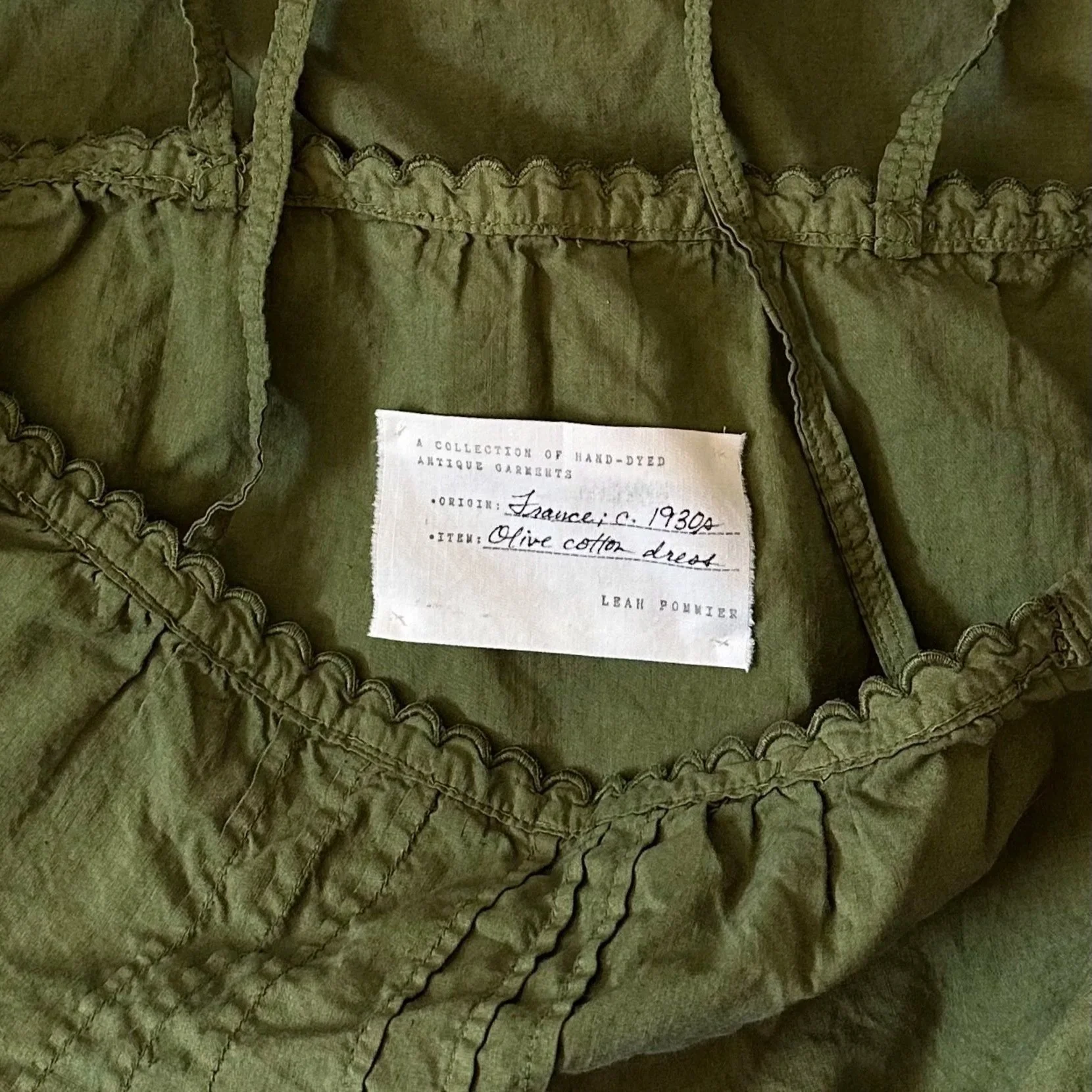 Close-up of a vintage olive-green garment with ruffled trim and a fabric label indicating it is a hand-dyed antique garment, originating from France around 1930, made of olive cotton.