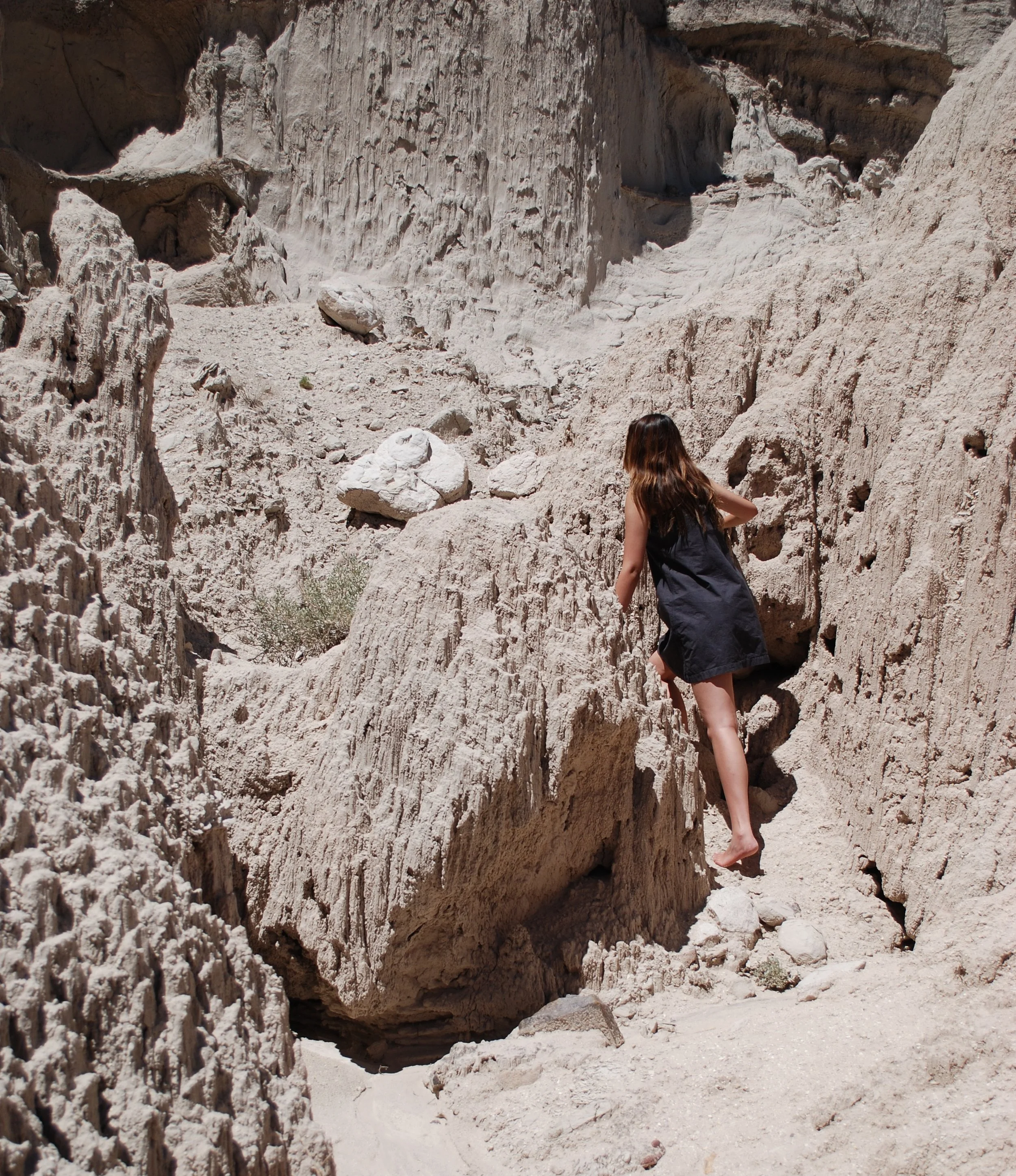 Woman with long hair and black dress climbing up a rocky, desert canyon with white sandstone walls.
