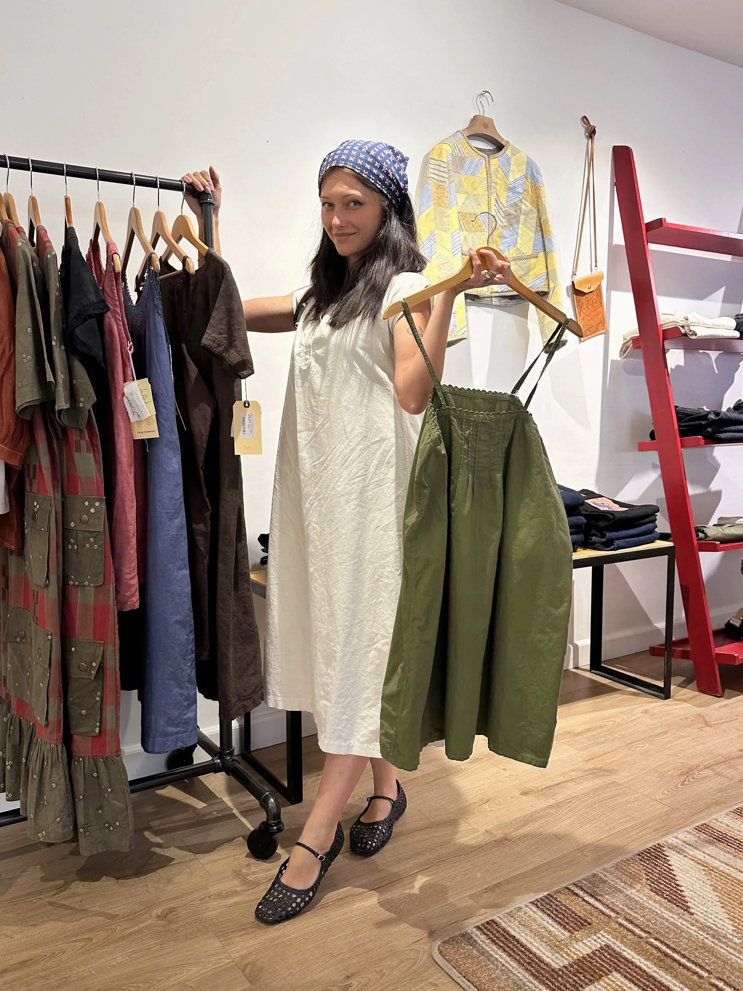 A woman in a white dress with a colorful headscarf and black flats holds a hanger with a green dress inside a clothing store.