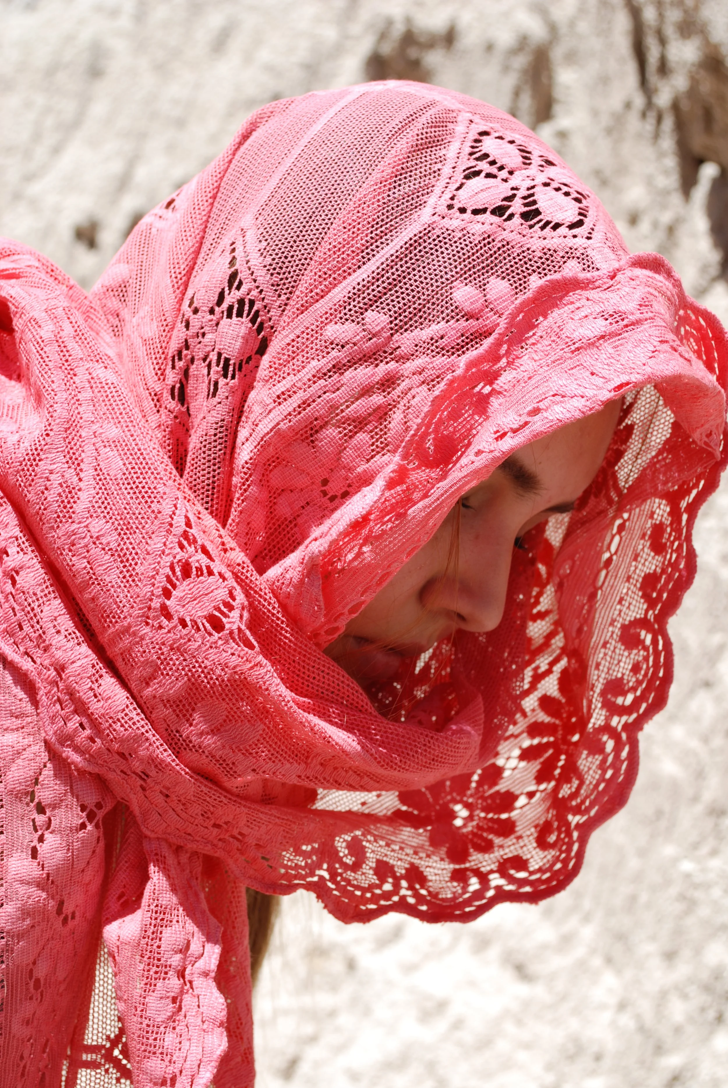 A person with fair skin wearing a pink lacy headscarf, with their face partially visible, looking downward, against a blurred sandy or rocky background.