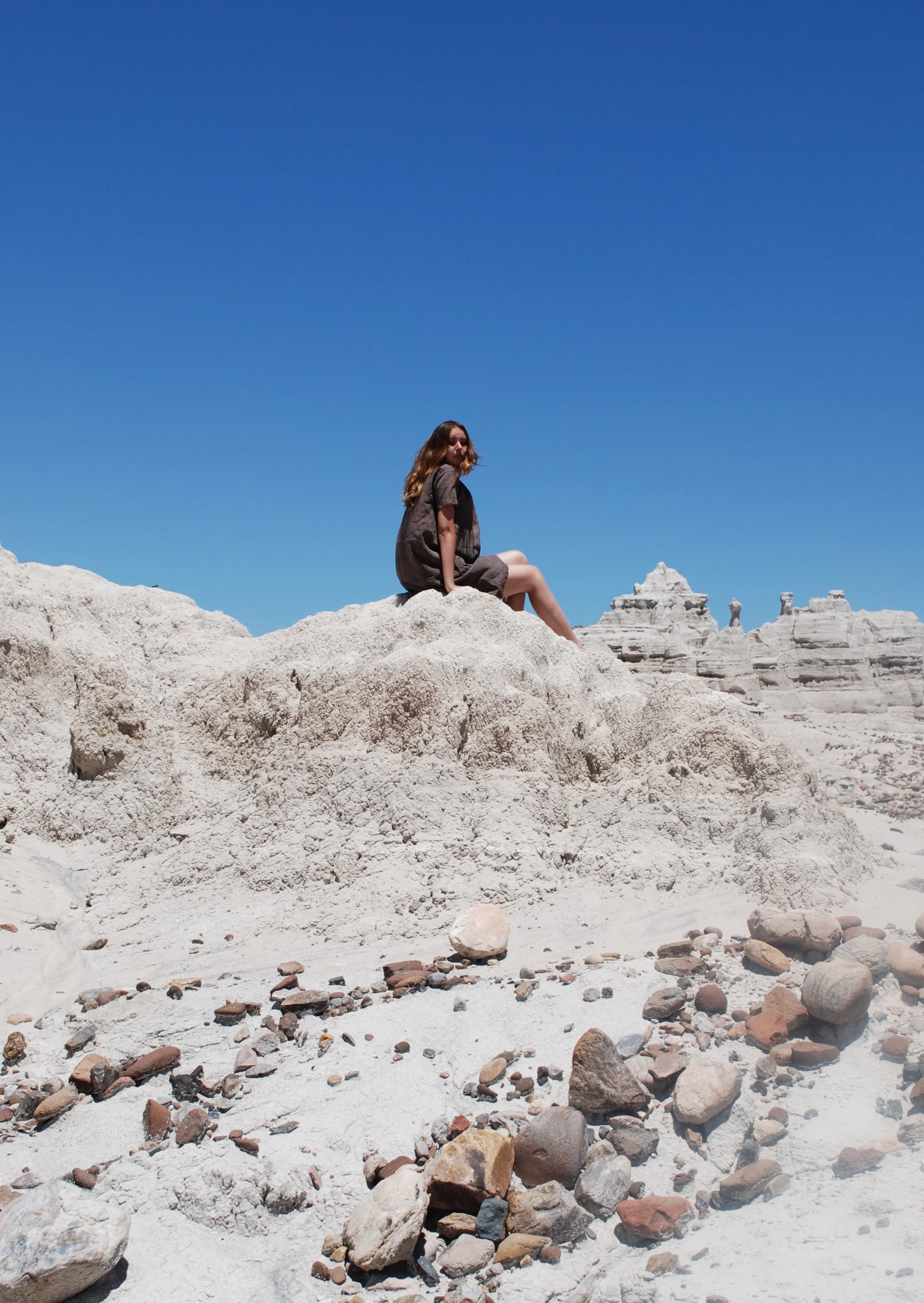 A young woman with long hair, wearing a dark dress, sitting on large white rocks in a desert landscape with white rock formations in the background and a clear blue sky above.