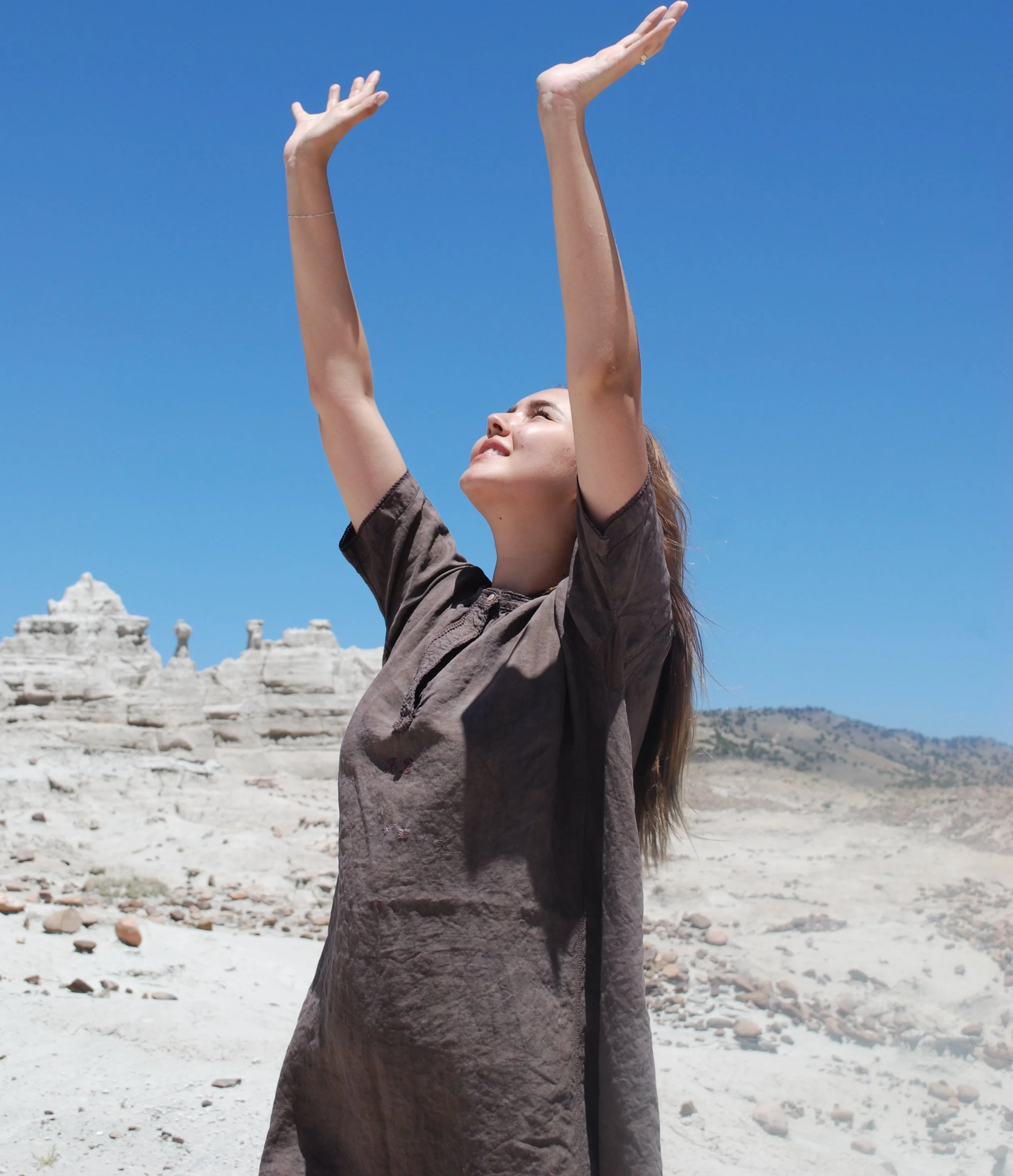 A woman with raised arms standing outdoors on a sunny day, with white ruins and mountains in the background.