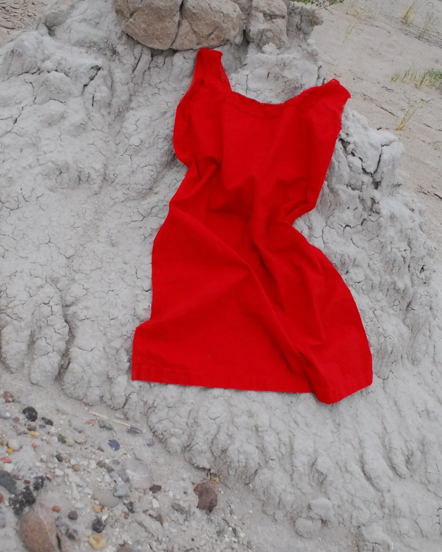 Red cloth or garment draped over cracked and uneven soil or ground with small rocks and sparse grass in the background.