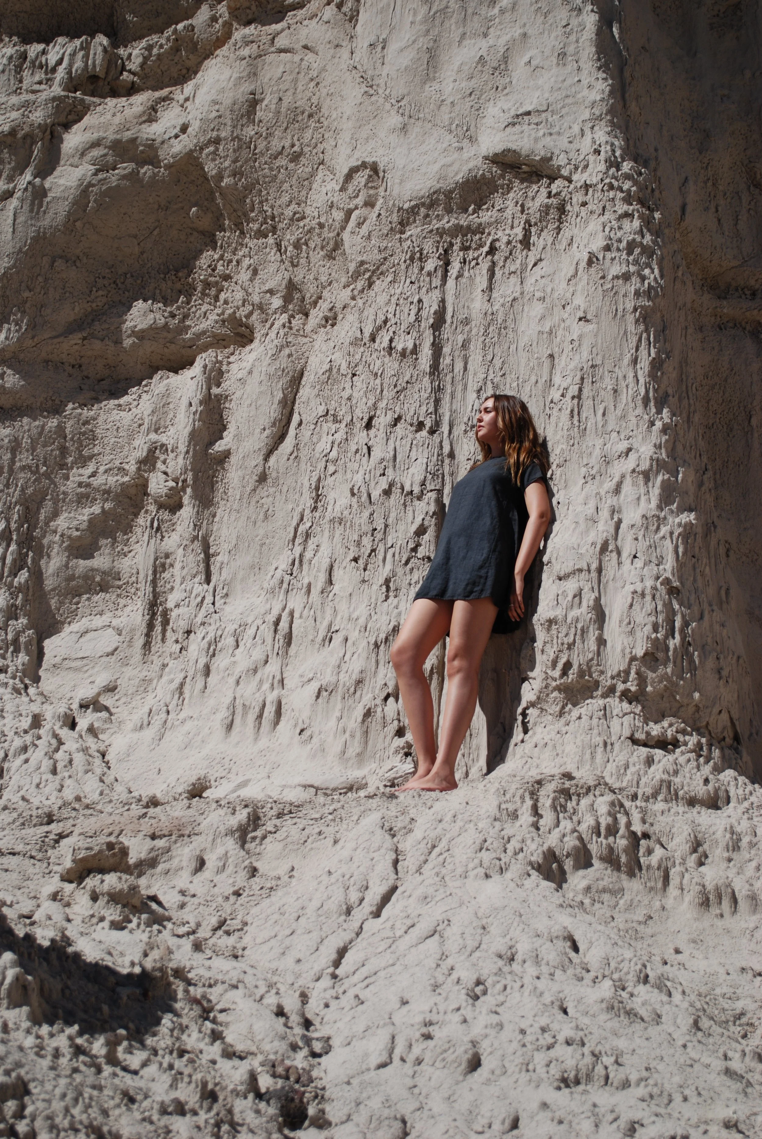 A woman with brown hair and a dark dress standing against a large white rock formation, with her eyes closed and face tilted upward. She is barefoot and the scene is outdoors, with sunlight illuminating the rocky backdrop.