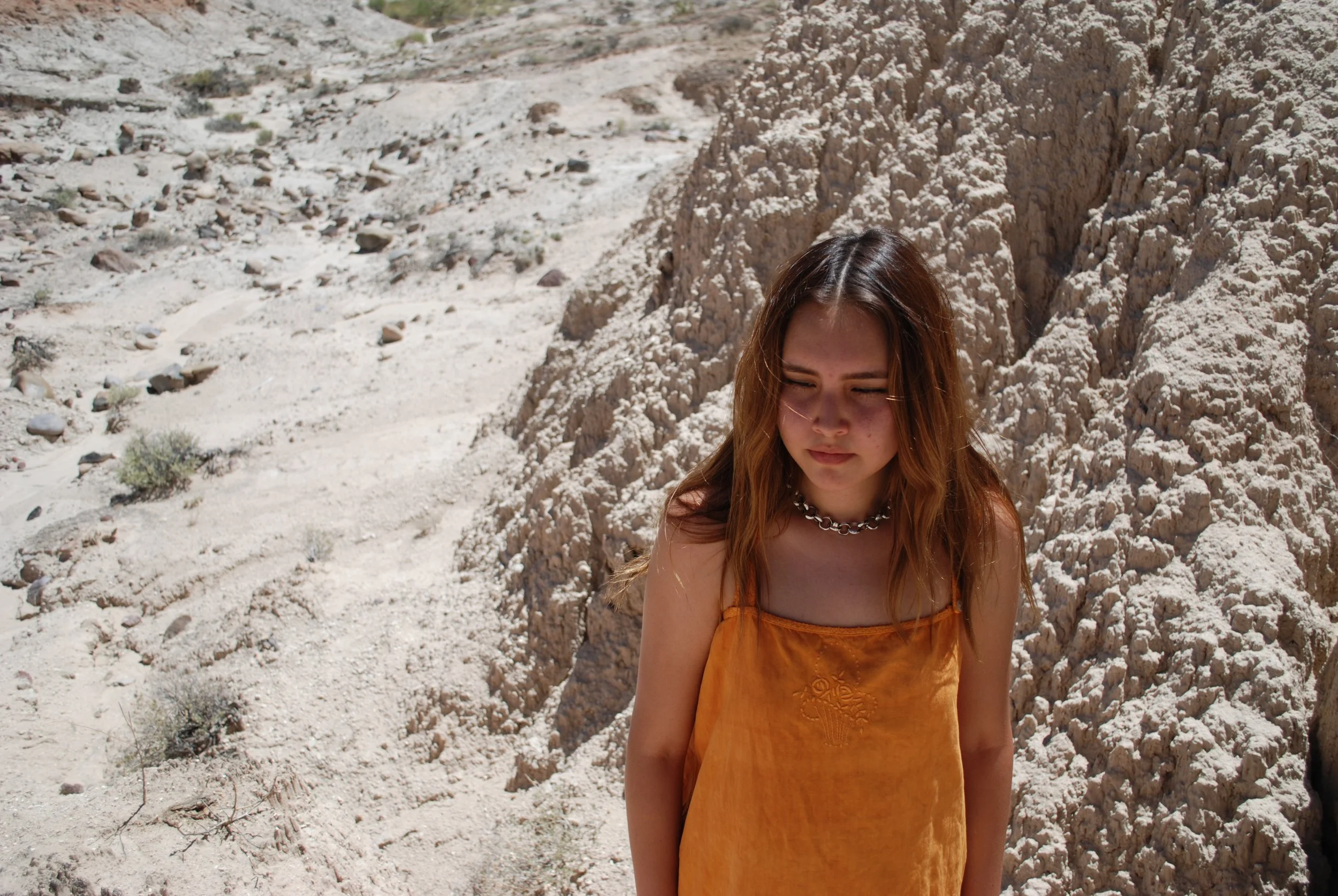 A young woman with long brown hair wearing an orange dress with embroidery, standing outdoors in a rocky, desert-like landscape with white, rugged terrain and sparse small bushes.