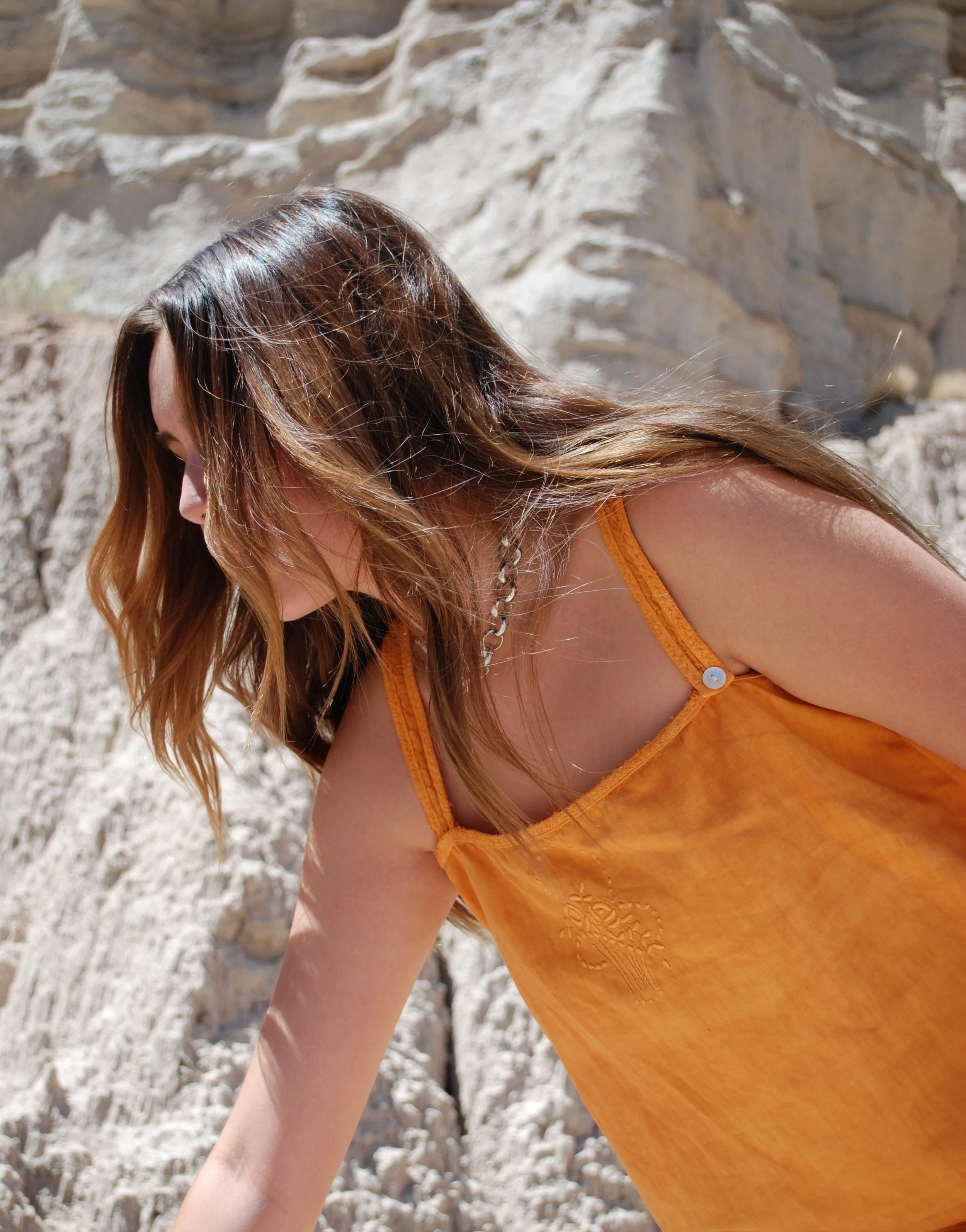 A woman with long, wavy brown hair wearing an orange top with embroidery, leaning forward with her head down, in front of a rocky background.