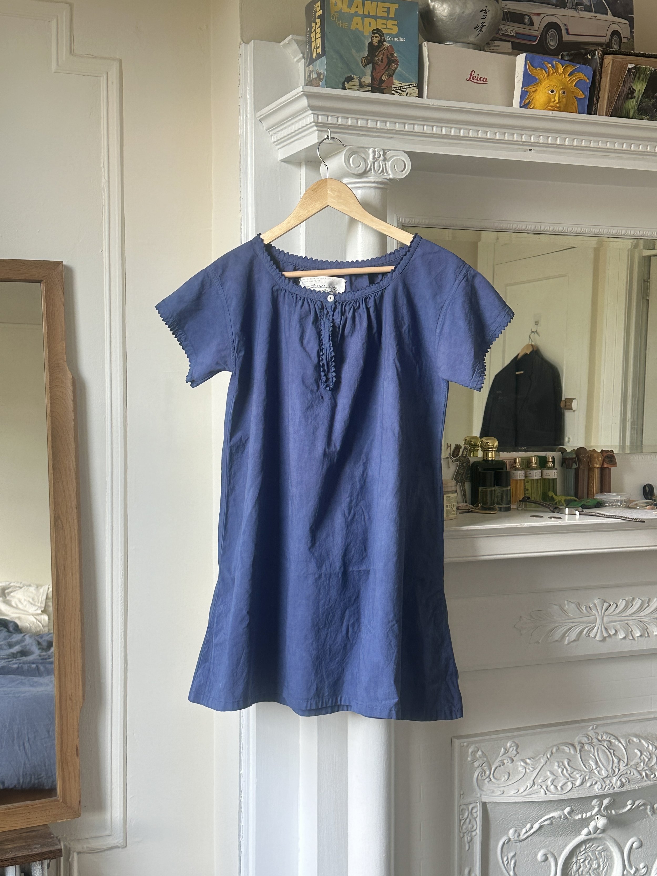 A blue dress with a gathered neckline hangs on a wooden hanger in front of a white ornate fireplace. Above, on the mantel, there are books and boxes, including a book titled 'Planet of the Apes,' with a box showing a car and other miscellaneous objects.