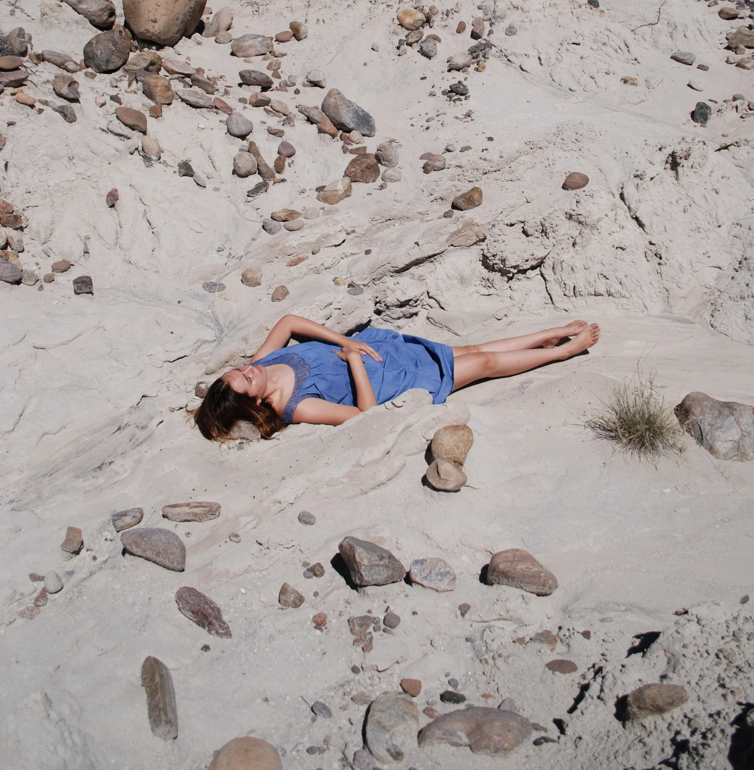A woman lying on a rocky, sandy beach with her eyes closed, wearing a blue dress.