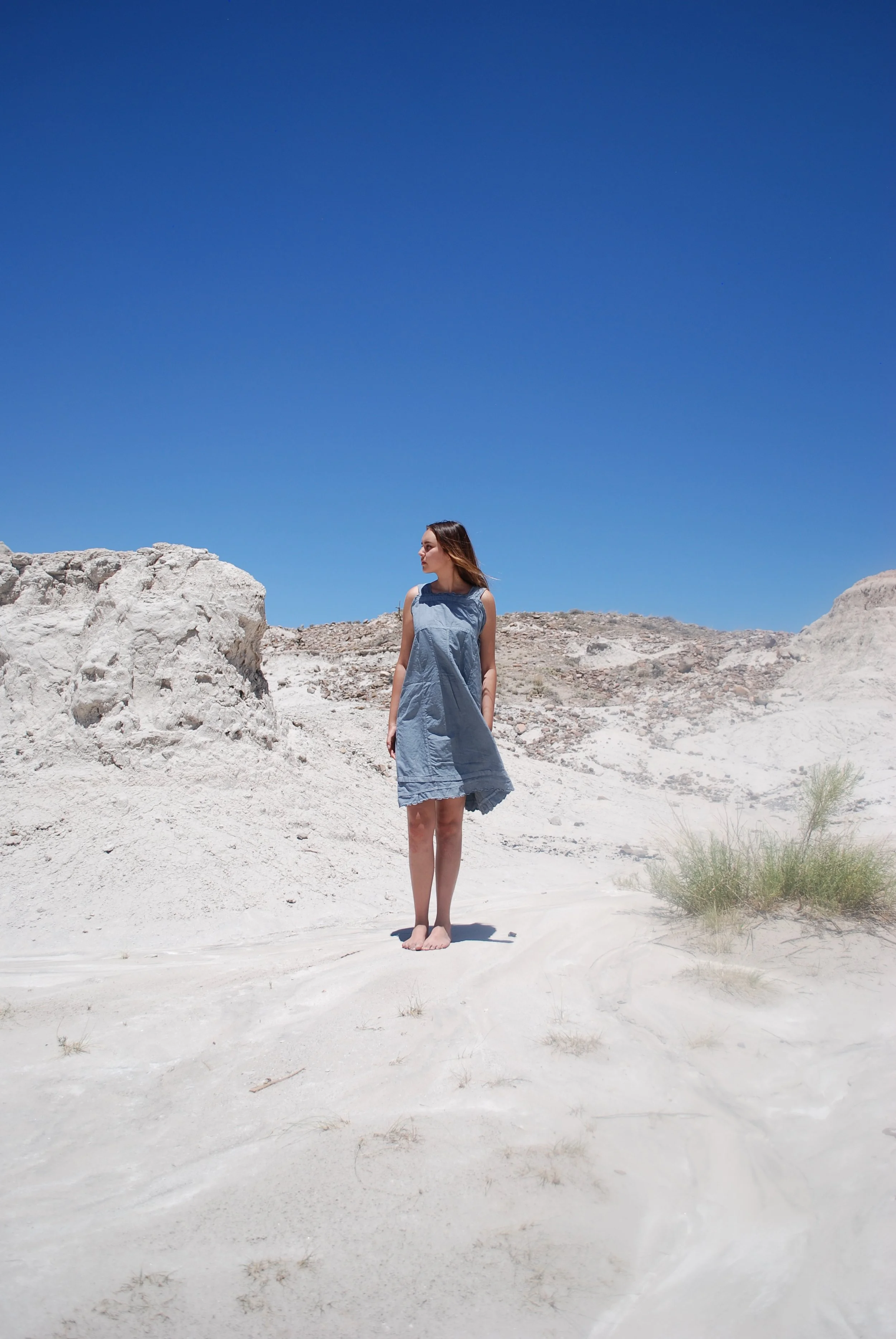 A woman with long hair in a sleeveless dress standing barefoot in a dry, rocky, desert-like landscape with white rocks and sparse vegetation under a clear blue sky.