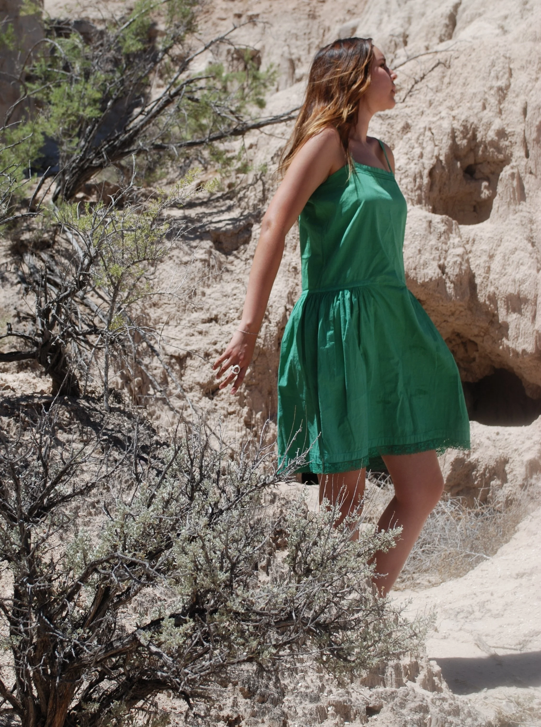 A woman wearing a green dress standing in a dry, rocky landscape with sparse vegetation and a large rock formation in the background.