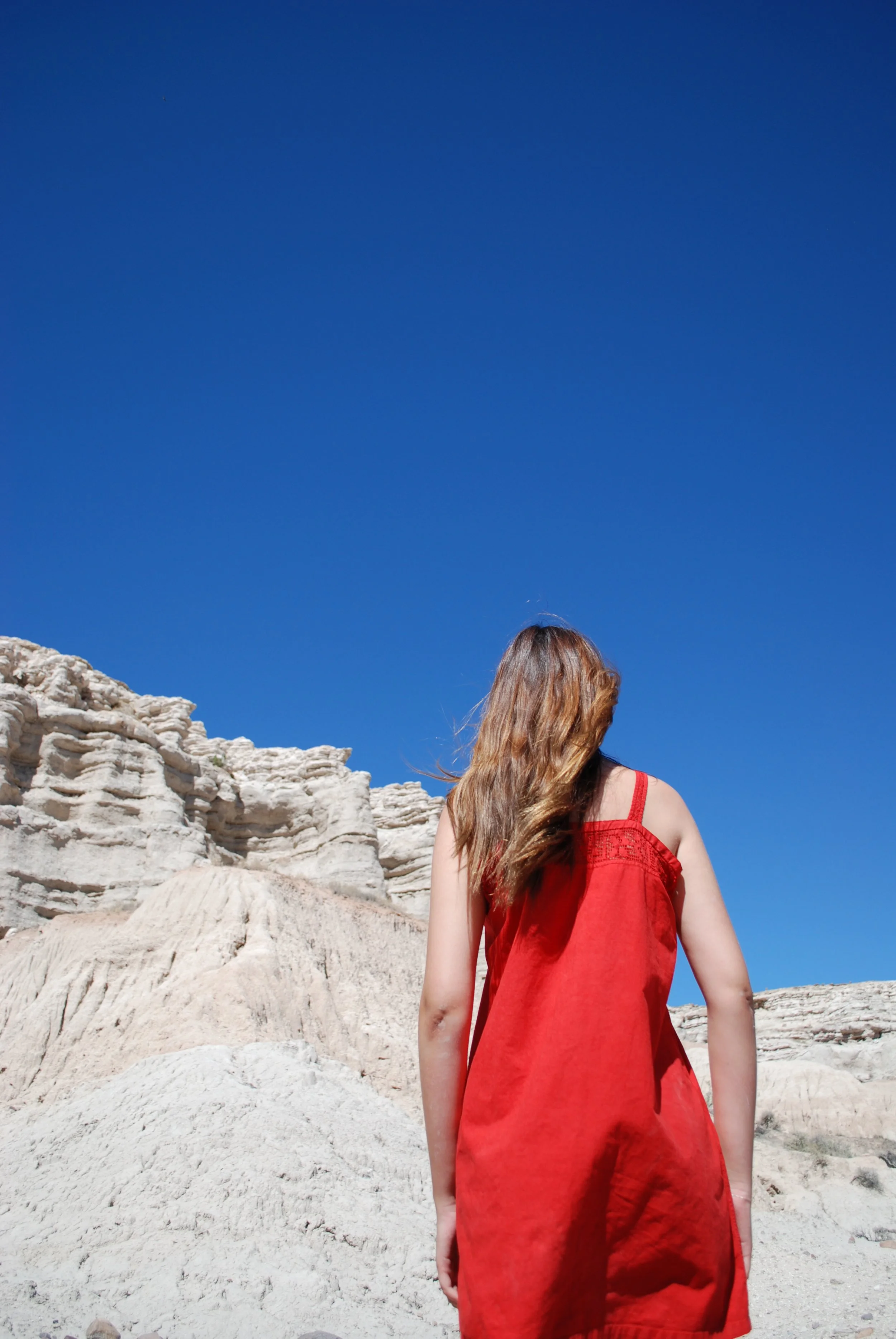 A woman with long, wavy hair wearing a sleeveless red dress standing outdoors against a backdrop of white rocky formations under a clear blue sky.