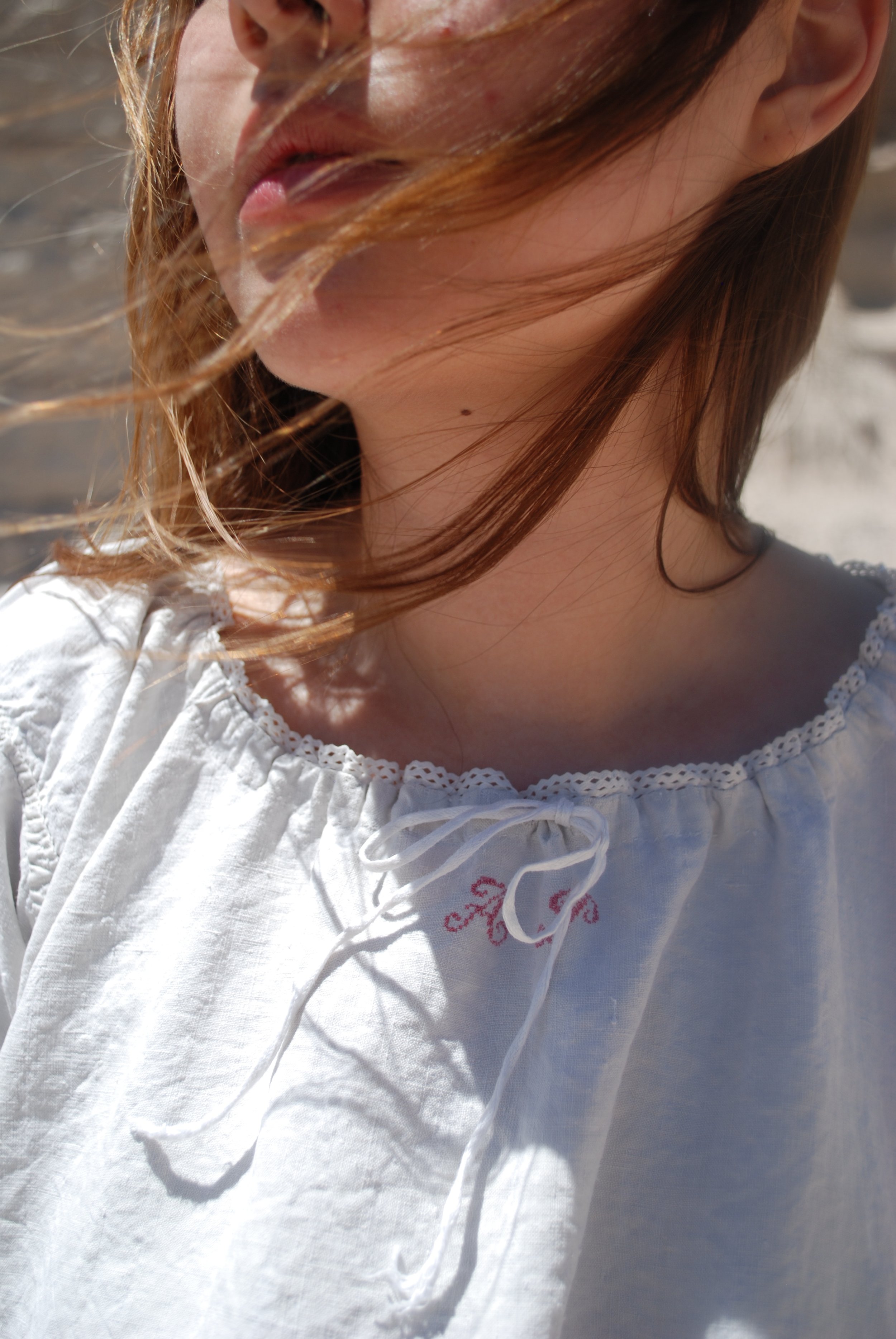 Close-up of a woman with reddish-brown hair, partly covered by windblown hair strands, wearing a white blouse with lace details and pink embroidery, outdoors in bright sunlight.