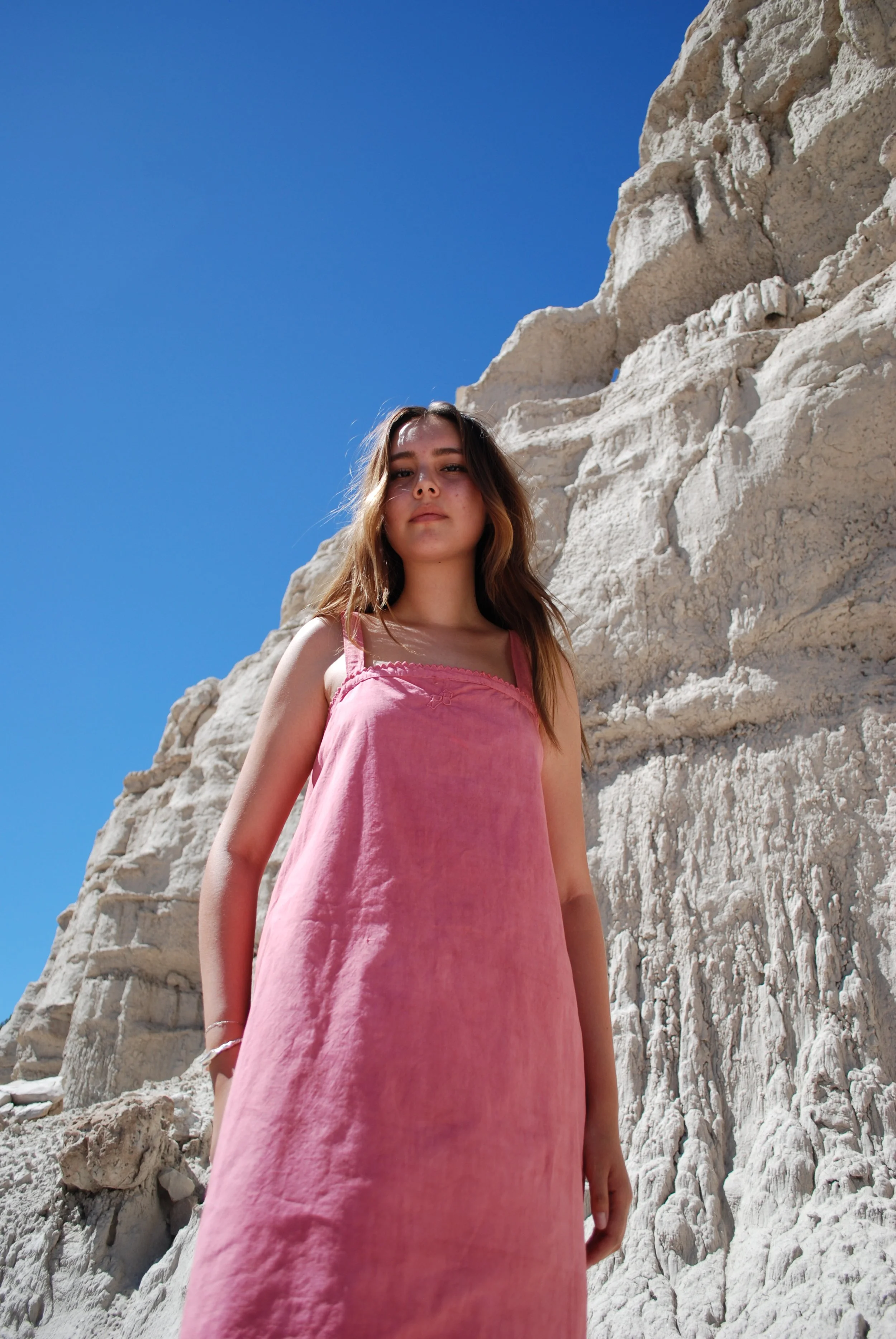 A young woman in a pink dress standing near white rocky cliffs under a clear blue sky.